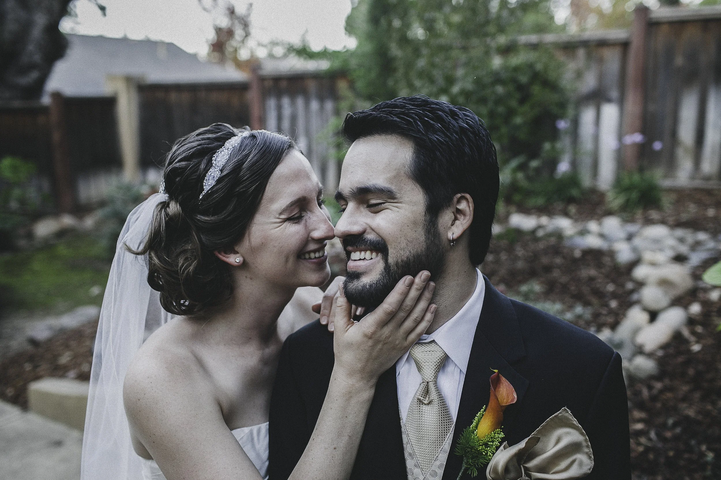 A bride and groom laugh and look at each other closely outside in a garden, with the bride touching the groom's face.