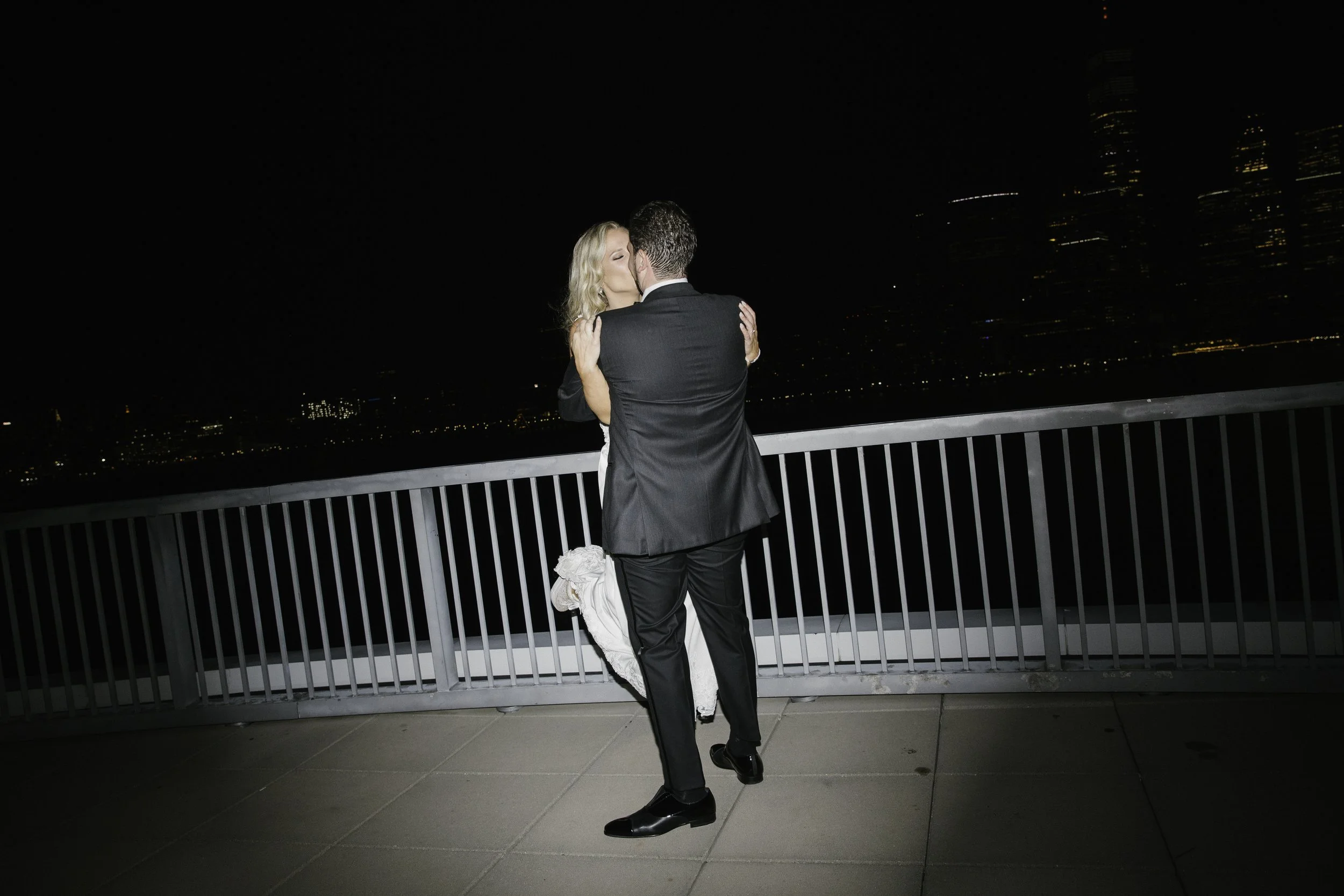 A couple dressed in wedding attire sharing a kiss on a rooftop at night with city skyline in the background.