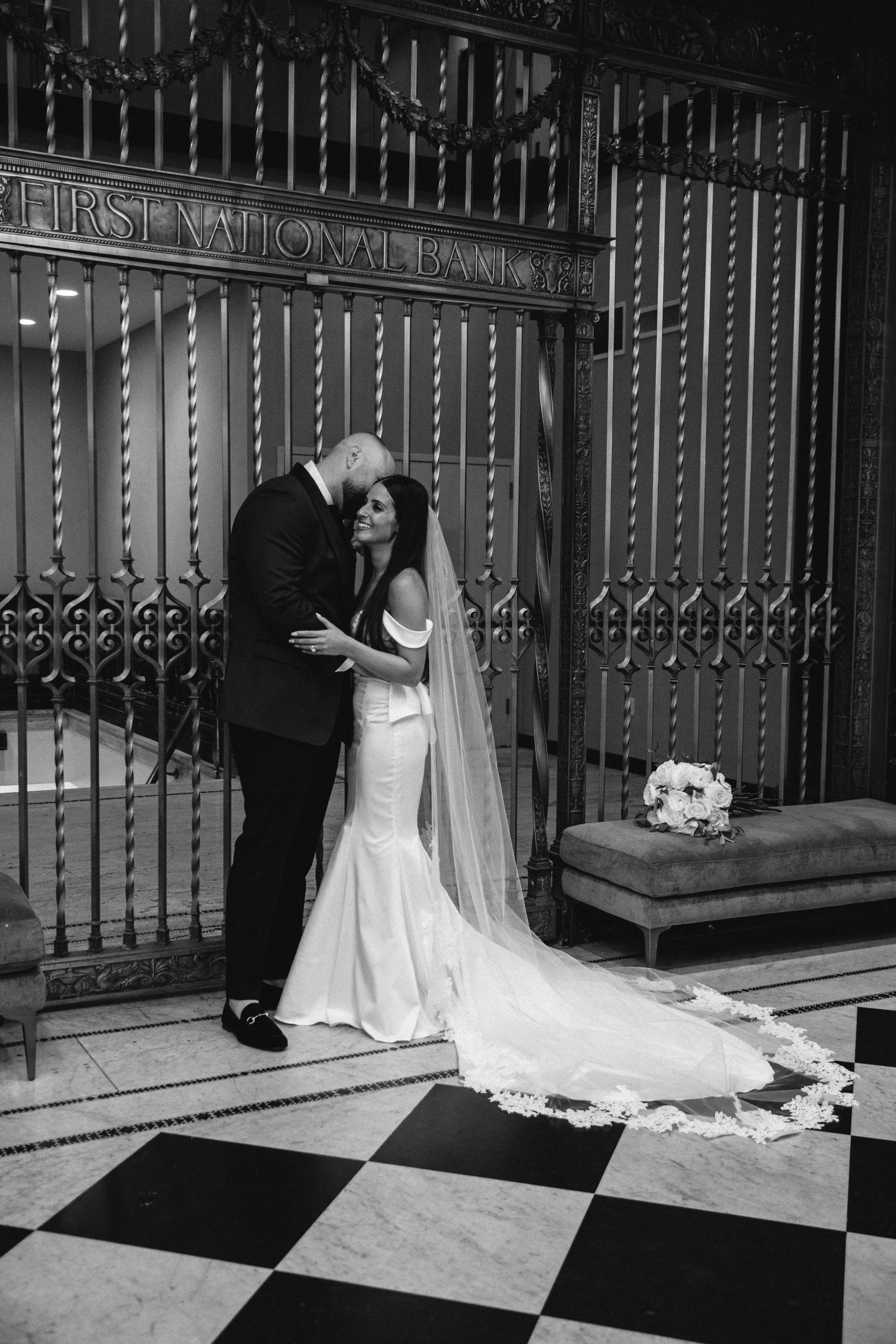 Black and white photo of a bride and groom embracing in front of a gated entrance, with the sign 'First National Bank' above them. The bride is wearing a wedding gown with a long train and veil, holding a bouquet of roses on a bench nearby. They are 