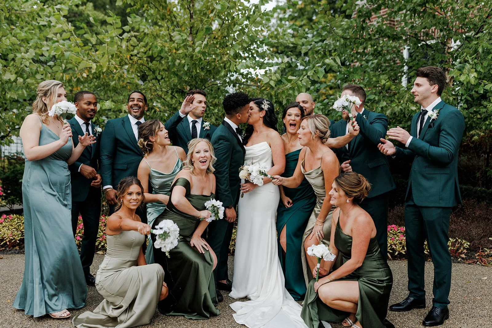 A group of wedding party members, including the bride and groom, celebrating outdoors with joyful expressions and playful gestures against a green leafy backdrop.