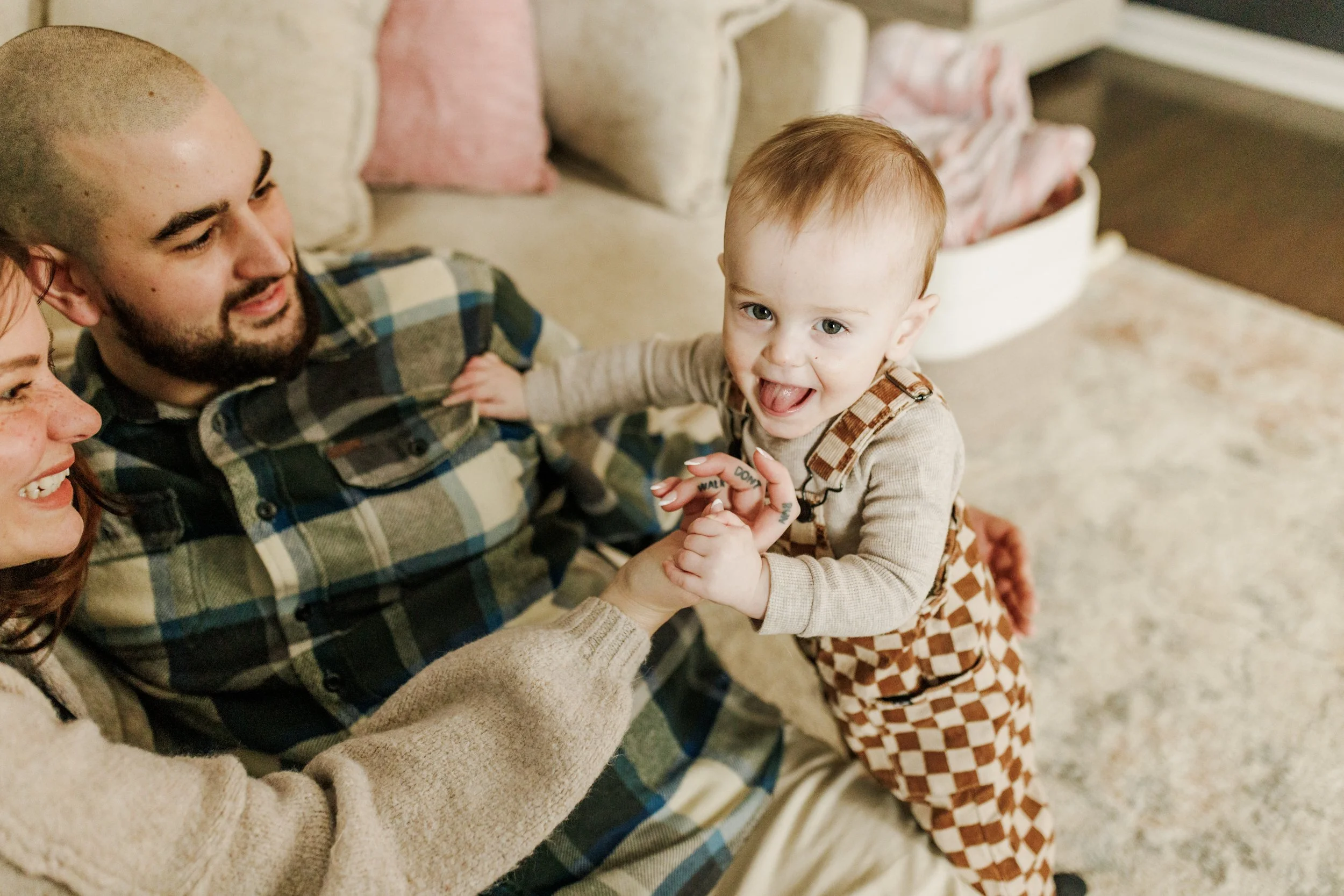 A young child is smiling and looking at the camera while holding hands with a woman and sitting on a man's lap on a couch in a cozy living room.