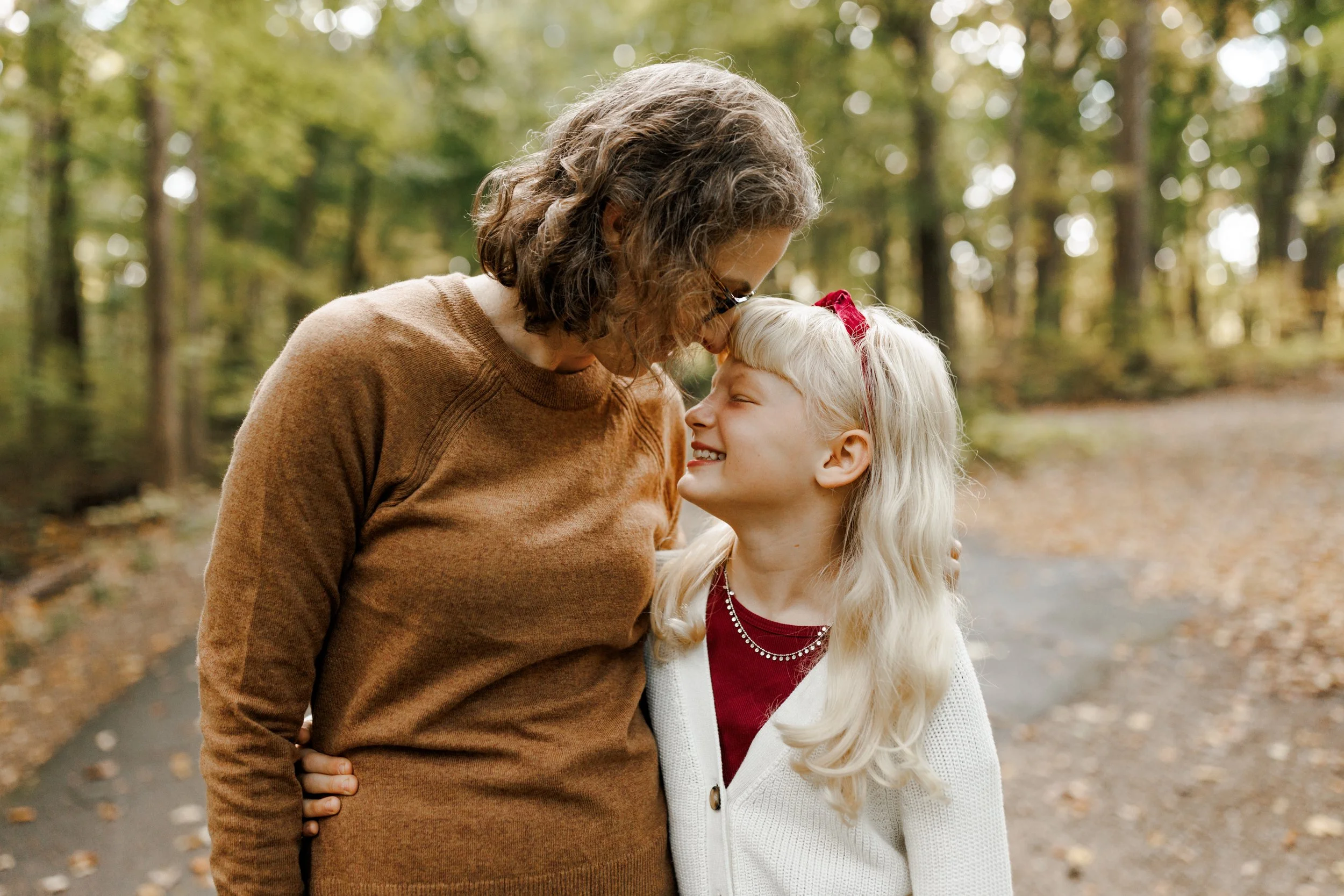 An elderly woman and a young girl sharing a joyful moment in a wooded park, touching foreheads and smiling at each other.