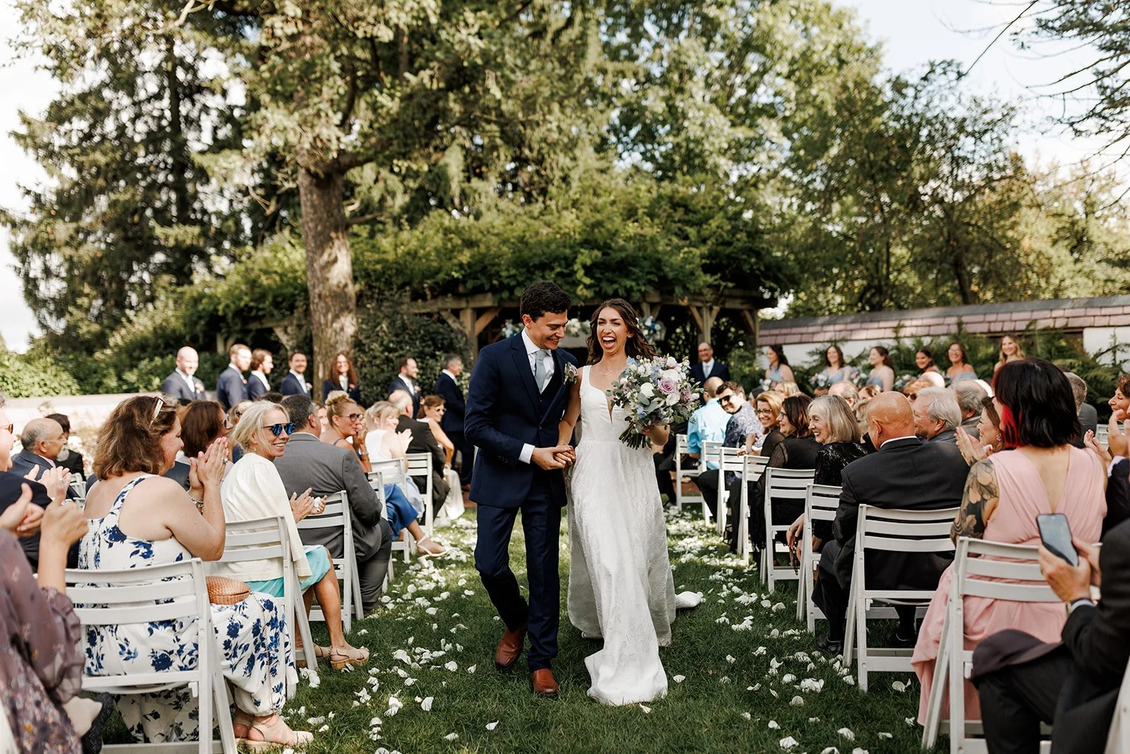 A bride and groom walking down the aisle, smiling and holding hands, during an outdoor wedding ceremony. The bride is wearing a white lace wedding dress and holding a bouquet of flowers. The groom is dressed in a dark suit with a white shirt and tie. Guests seated on white chairs are clapping and smiling, with some taking photos. The setting is a lush garden with green trees and a wooden arch in the background.