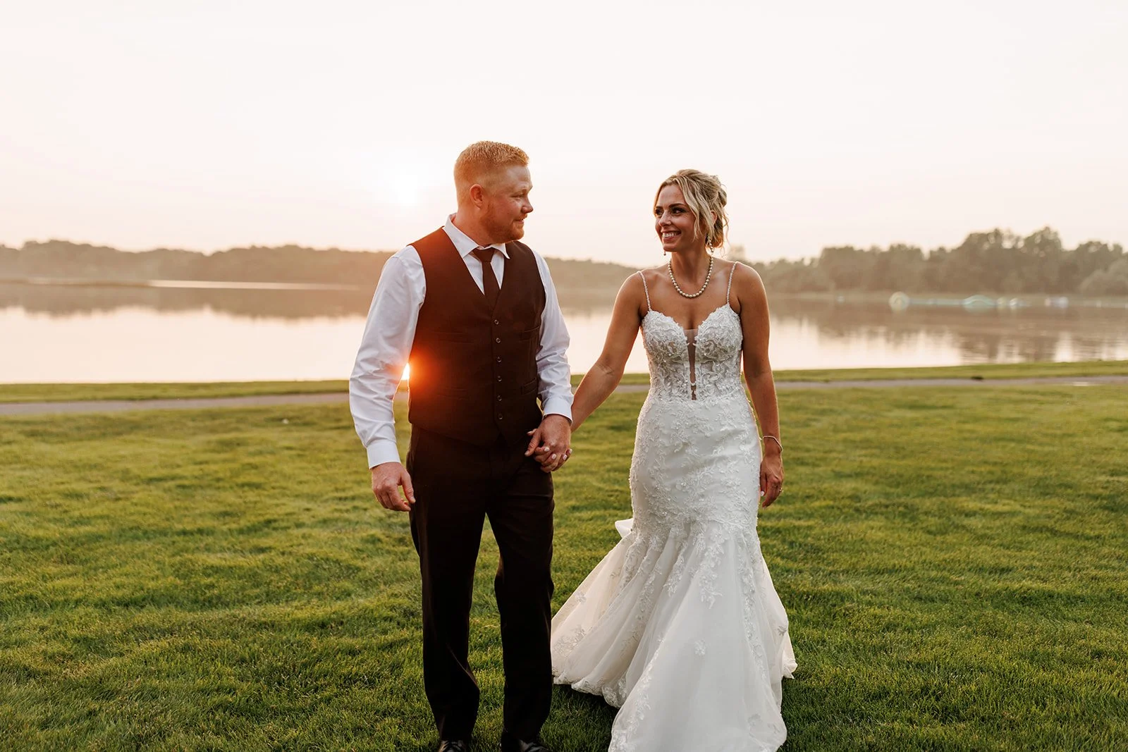 A newlywed couple walking hand in hand on a grassy area near a lake during sunset, with the bride wearing a lace wedding gown and the groom in a black vest and white shirt.