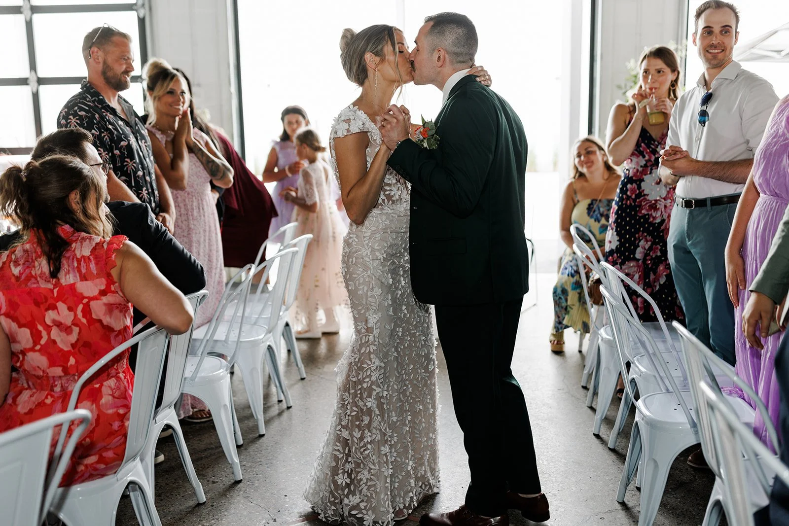 A bride and groom share a kiss while holding hands at their wedding ceremony, surrounded by guests in a bright venue with large windows.
