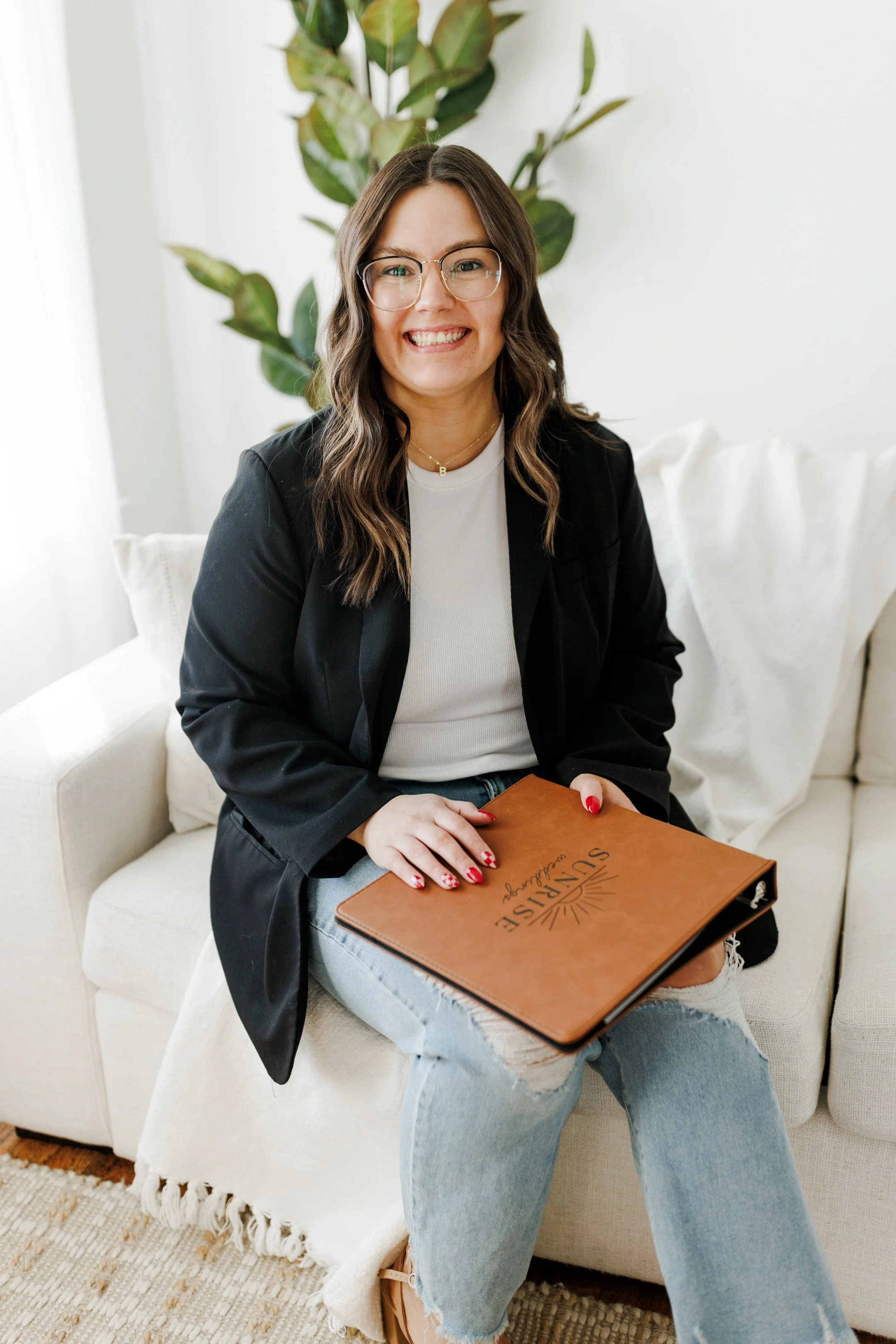 A woman with wavy brown hair, glasses, and light skin, sitting on a cream-colored sofa, smiling, holding a brown menu or folder that says 'Sunrise Washington' with a sun illustration, in a bright room with a plant in the background.