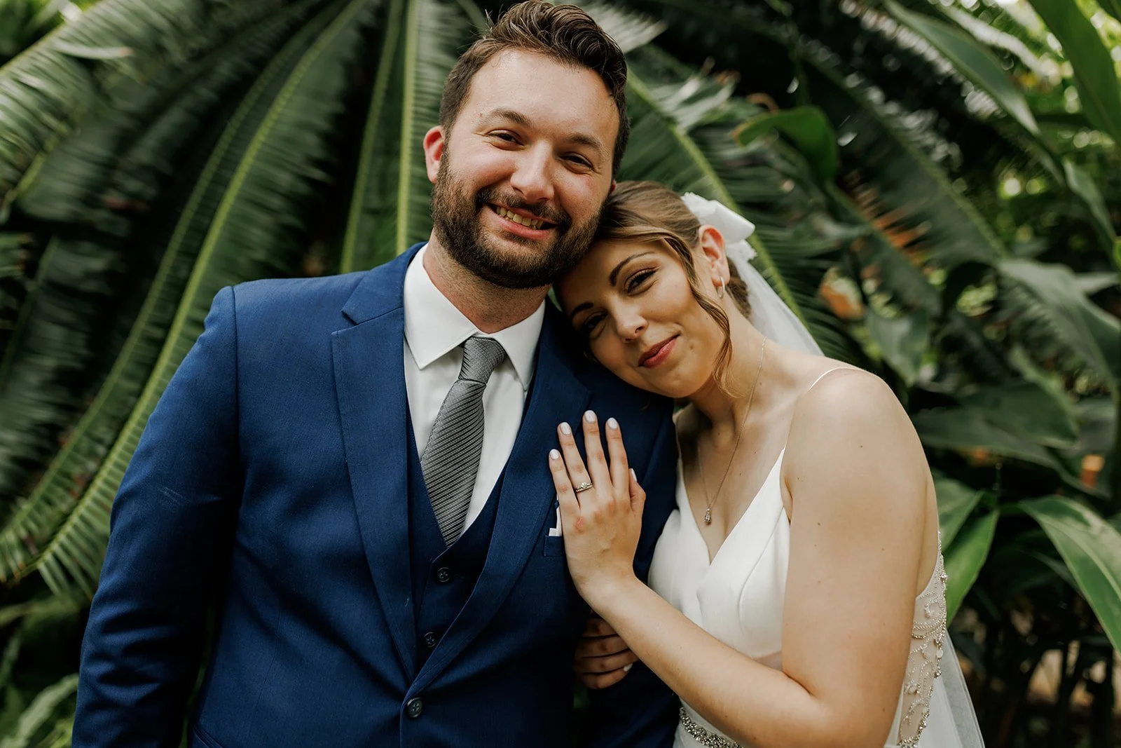 A newlywed couple smiling and posing outdoors with lush green tropical plants in the background, the bride resting her head on the groom's shoulder.