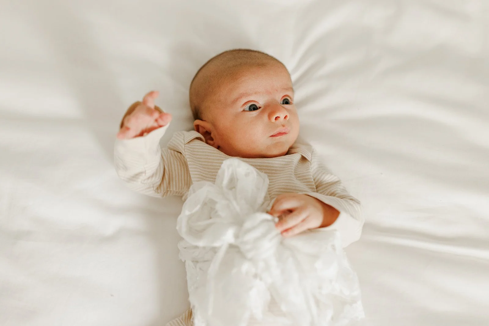 Baby lying on a white bed holding a crumpled paper, wearing a beige striped outfit, with wide eyes and a curious expression.