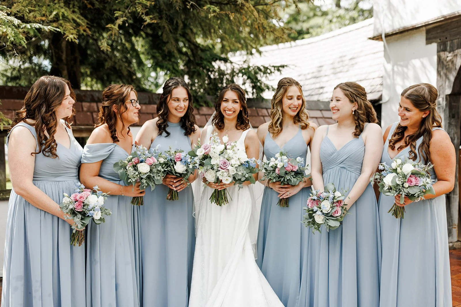 A bride in a white wedding dress surrounded by seven bridesmaids in light blue dresses, holding bouquets of flowers, standing outdoors with trees and a rustic building in the background.