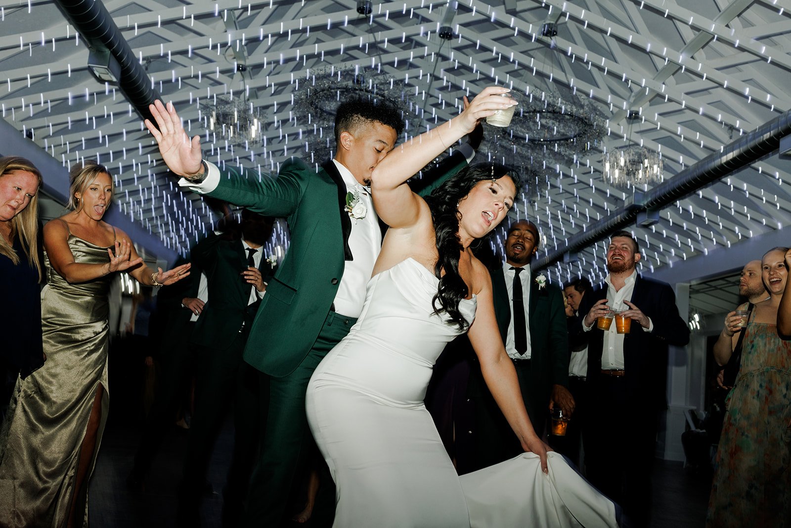 A bride and groom dancing at their wedding reception with friends and family surrounding them, under a ceiling decorated with string lights.