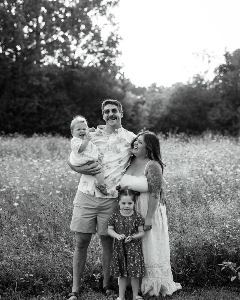 A family of four standing in a field of wildflowers, smiling and posing for a photo in black and white.