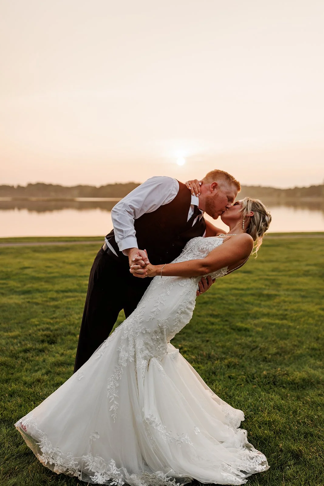 A newlywed couple sharing a kiss during sunset by a lake, with the groom dipping the bride. The bride is wearing a white wedding dress with lace details, and the groom is dressed in a black vest and white shirt. The scene is romantic and picturesque.