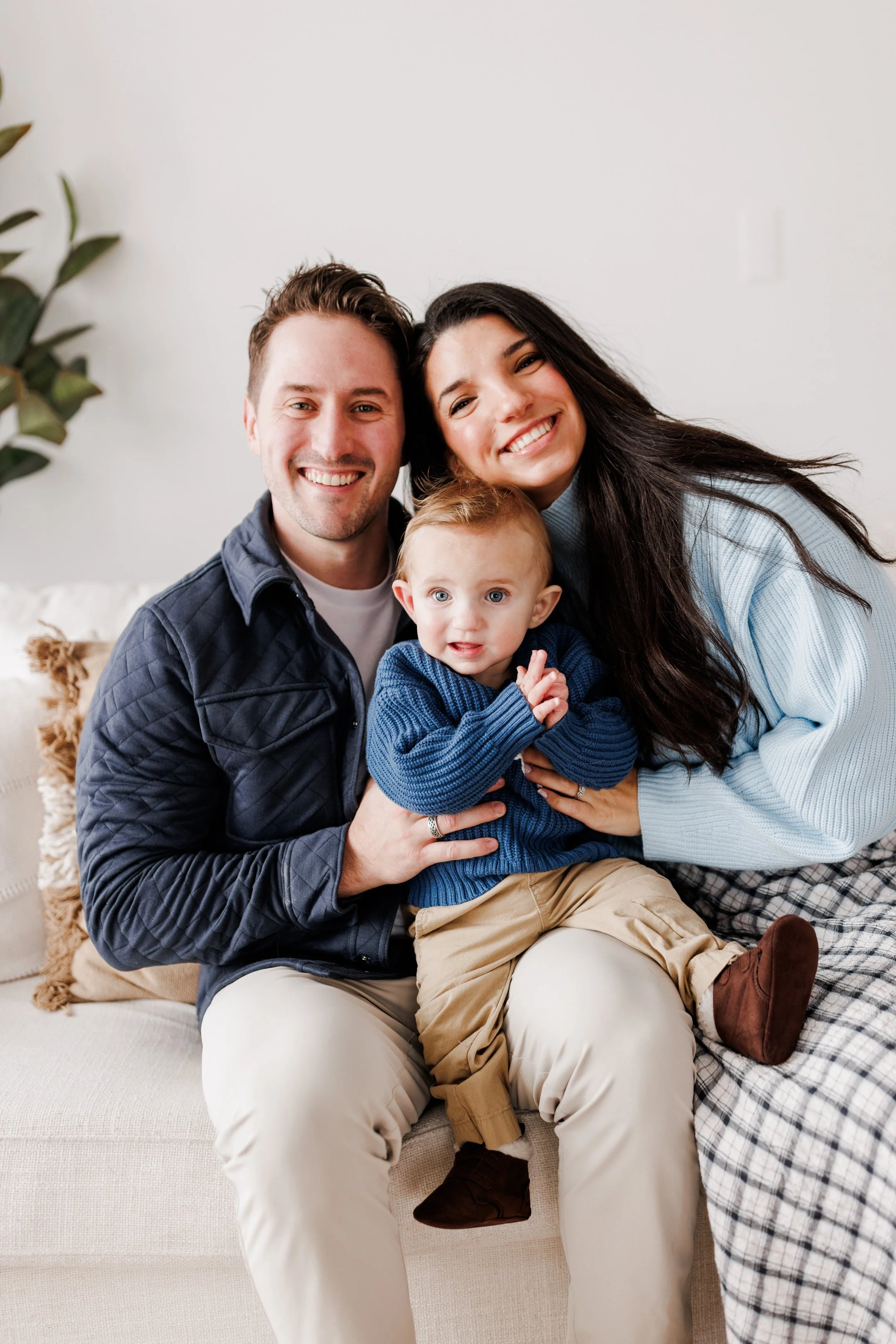 A happy family of three sitting on a beige sofa, smiling at the camera. The father has short brown hair and is wearing a dark quilted jacket. The mother has long dark hair and is wearing a light blue sweater. The young child has red hair, blue eyes, 
