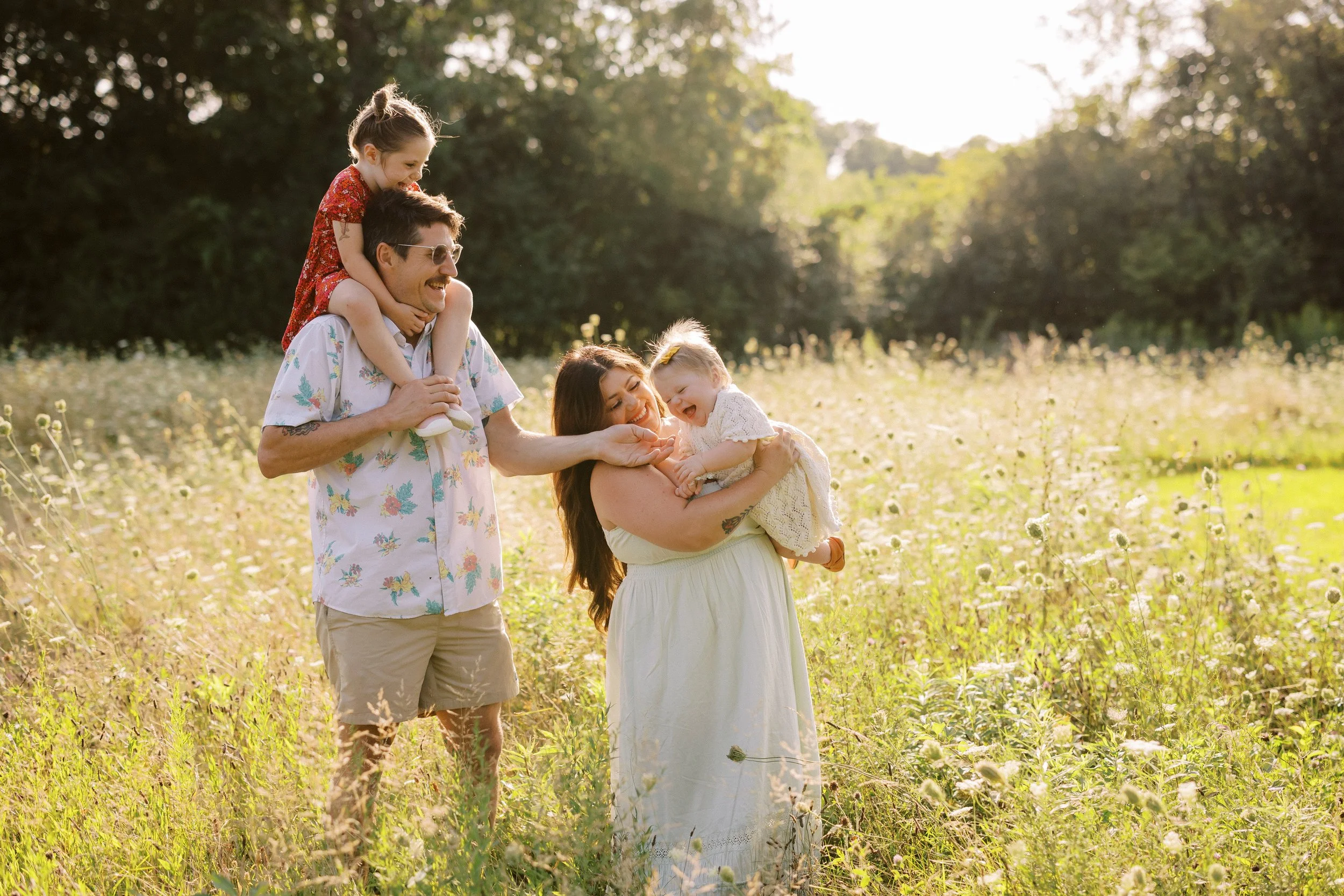 A happy family of four enjoying a sunny day in a grassy field with wildflowers, with children playing on the adults' shoulders and laughing.