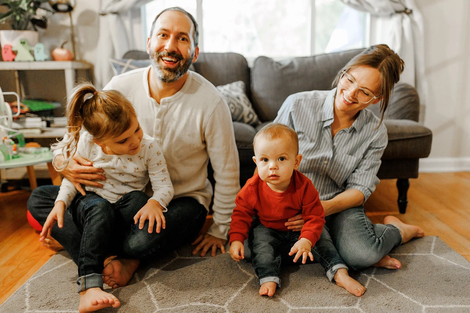 A happy family playing on the living room floor. There are two children, two adults, and a rug. The adults are smiling, and one of the children is looking at the camera.