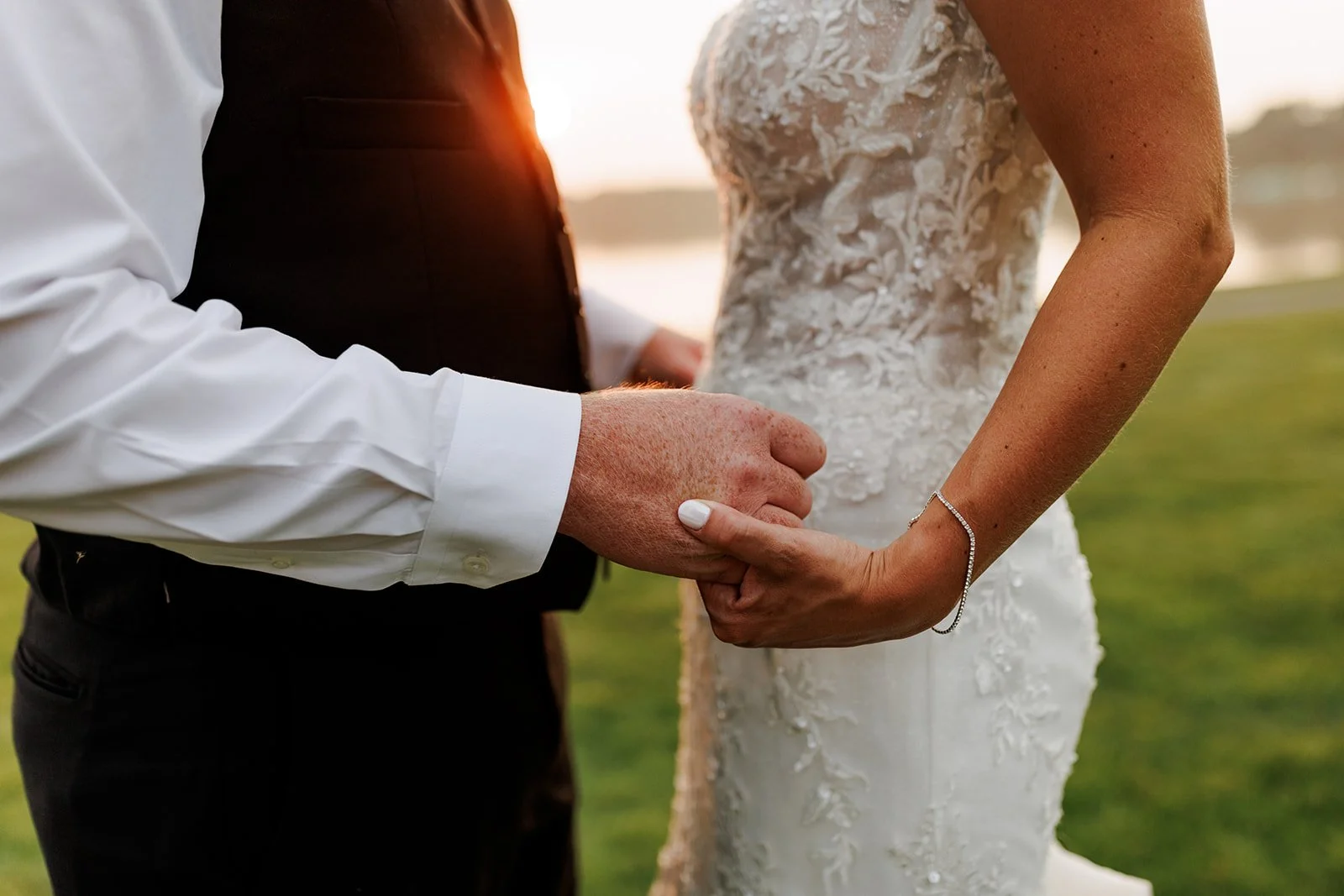 A couple holding hands during their wedding, with a blurred outdoor background and sunlight.
