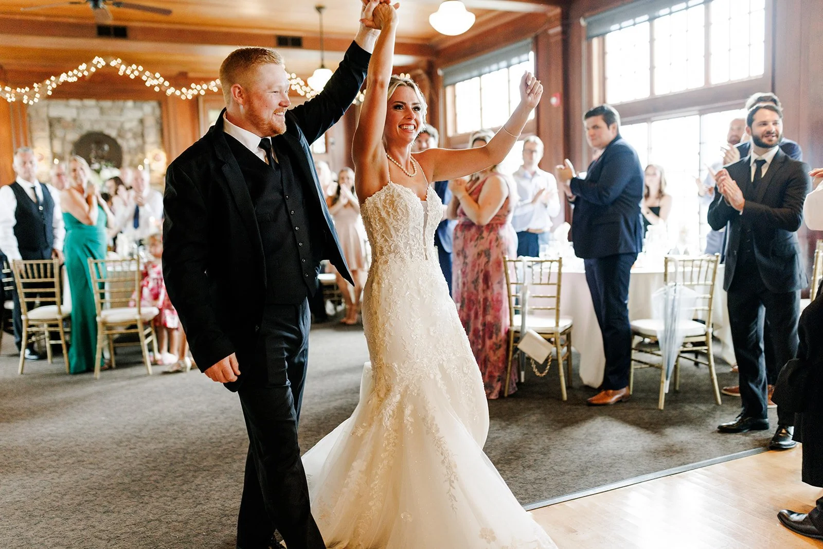 Bride and groom dancing hand in hand at wedding reception with guests clapping and celebrating inside a decorated venue.