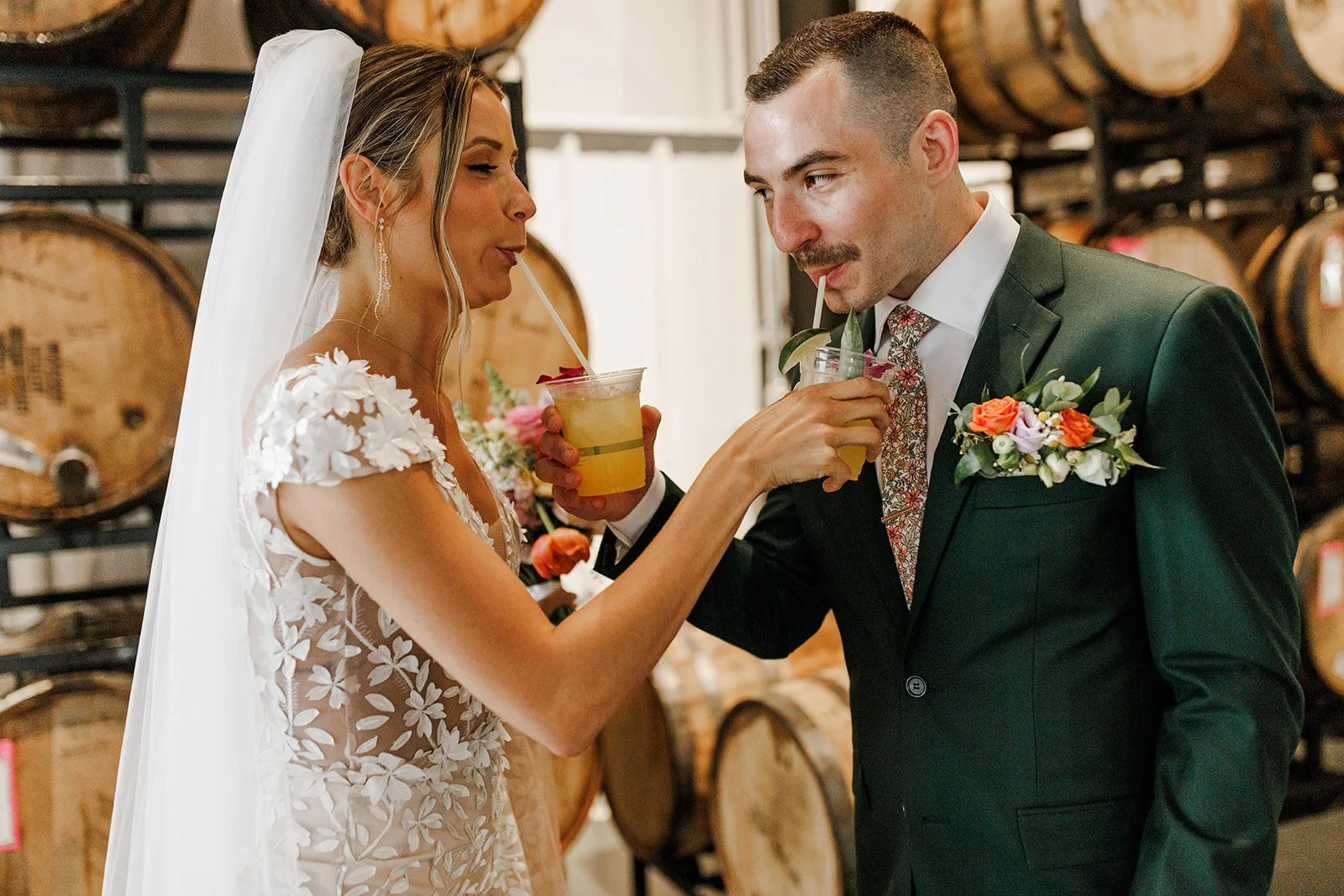 A bride and groom sharing a drink with straws at their wedding reception. The bride wears a lace wedding dress and a veil, and the groom wears a dark suit with a boutonnière. They are smiling and standing in front of wine barrels.