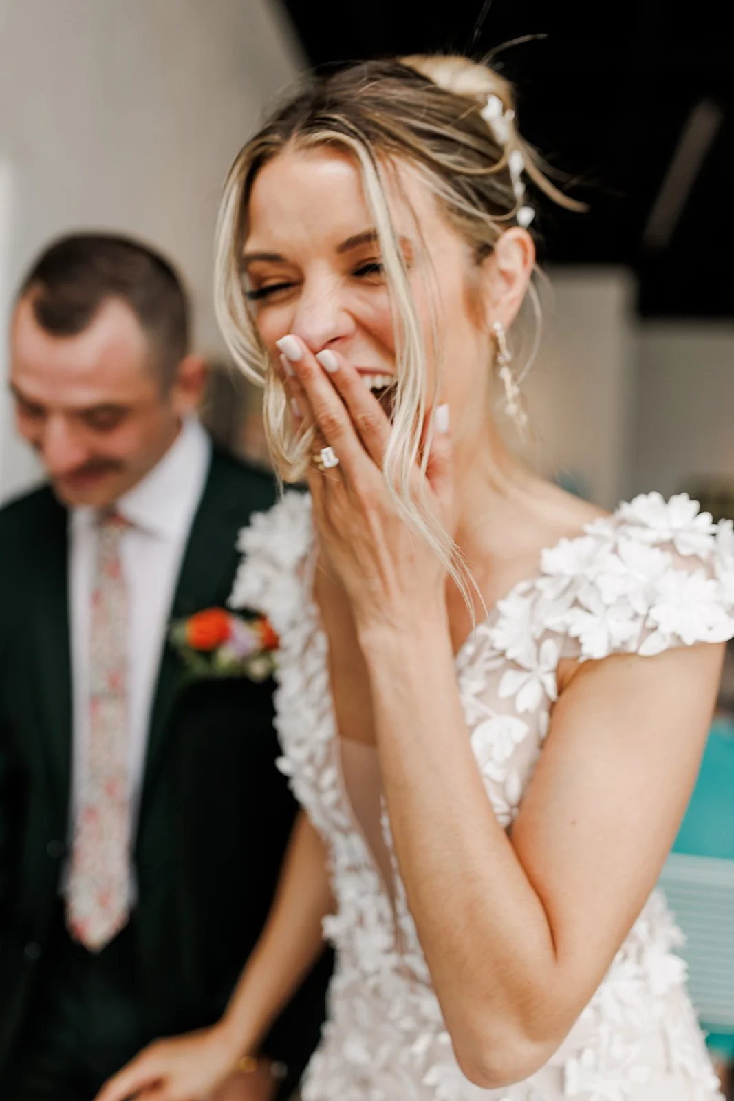 A bride laughing with her hand covering her mouth, wearing a white dress with floral appliqué, and a man in a suit with a boutonnière stands beside her, smiling.