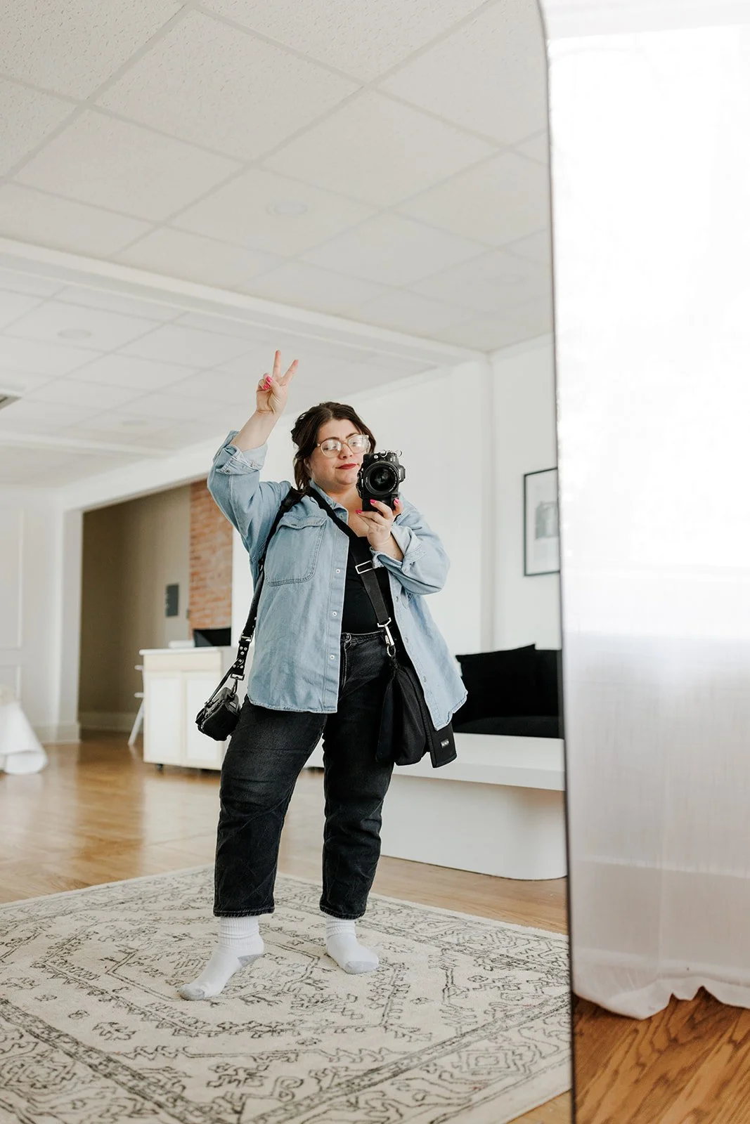 A woman taking a mirror selfie in a room, making a peace sign with her right hand, wearing glasses, a denim jacket, black pants, white socks, and carrying a camera.