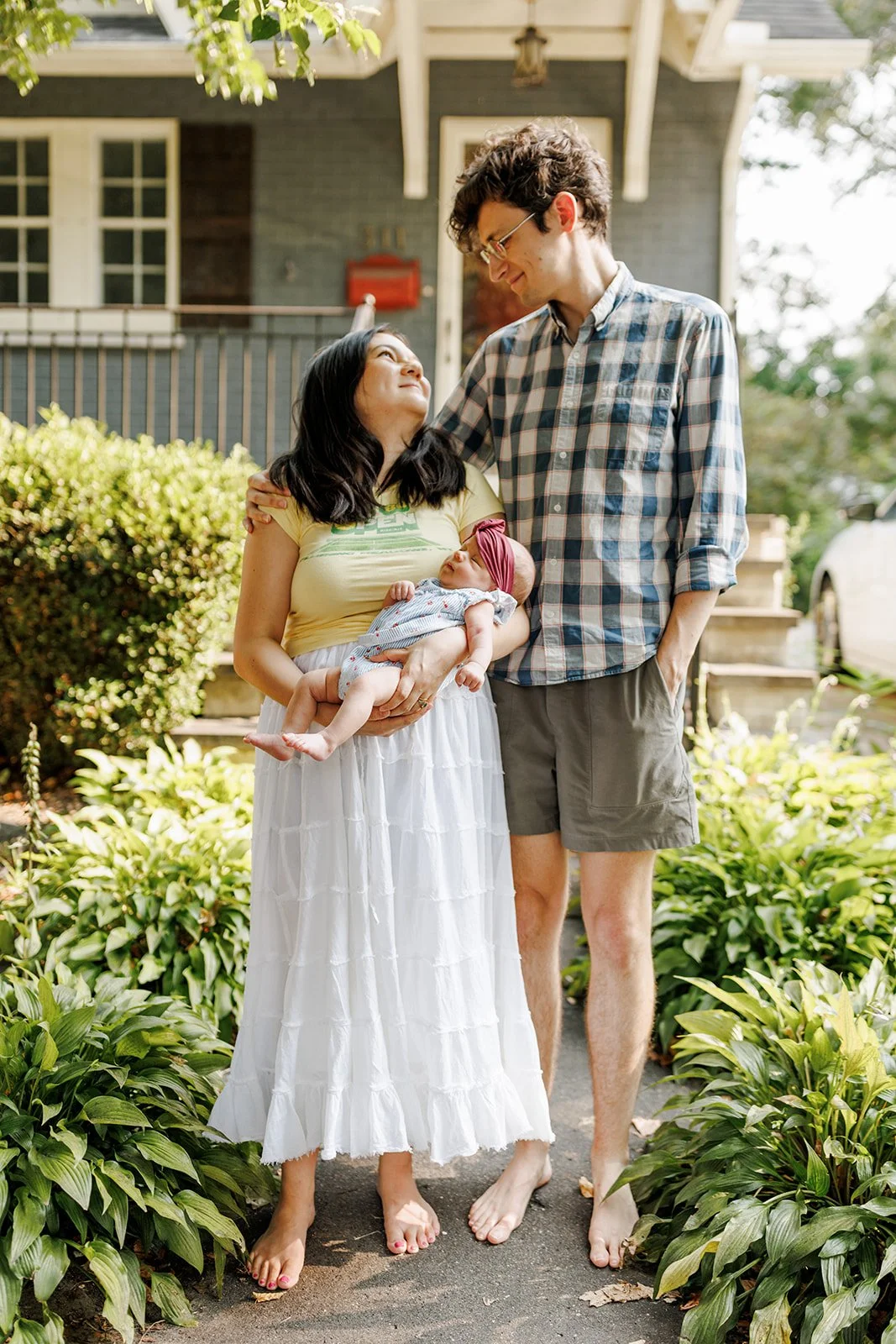 A woman holding a baby, standing next to a man outdoors near a house, on a sunny day.