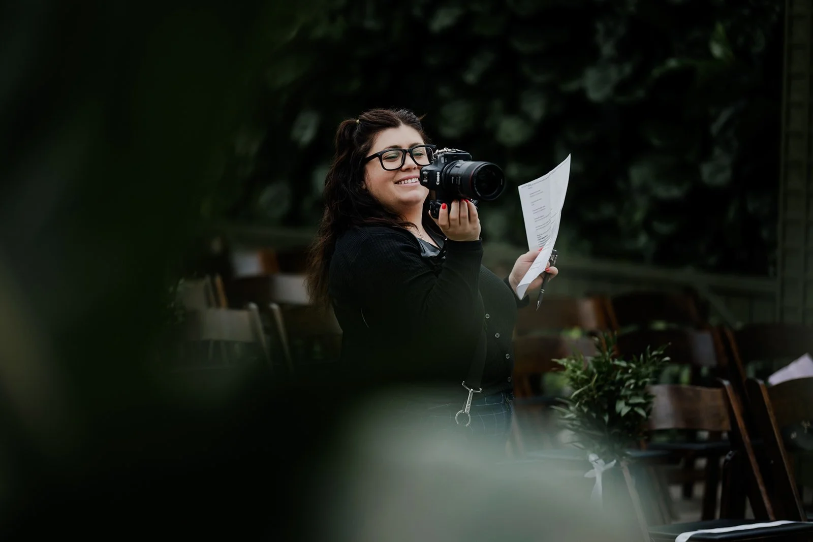 A young woman with glasses, holding a camera and a piece of paper, smiling in an outdoor setting surrounded by greenery and chairs.