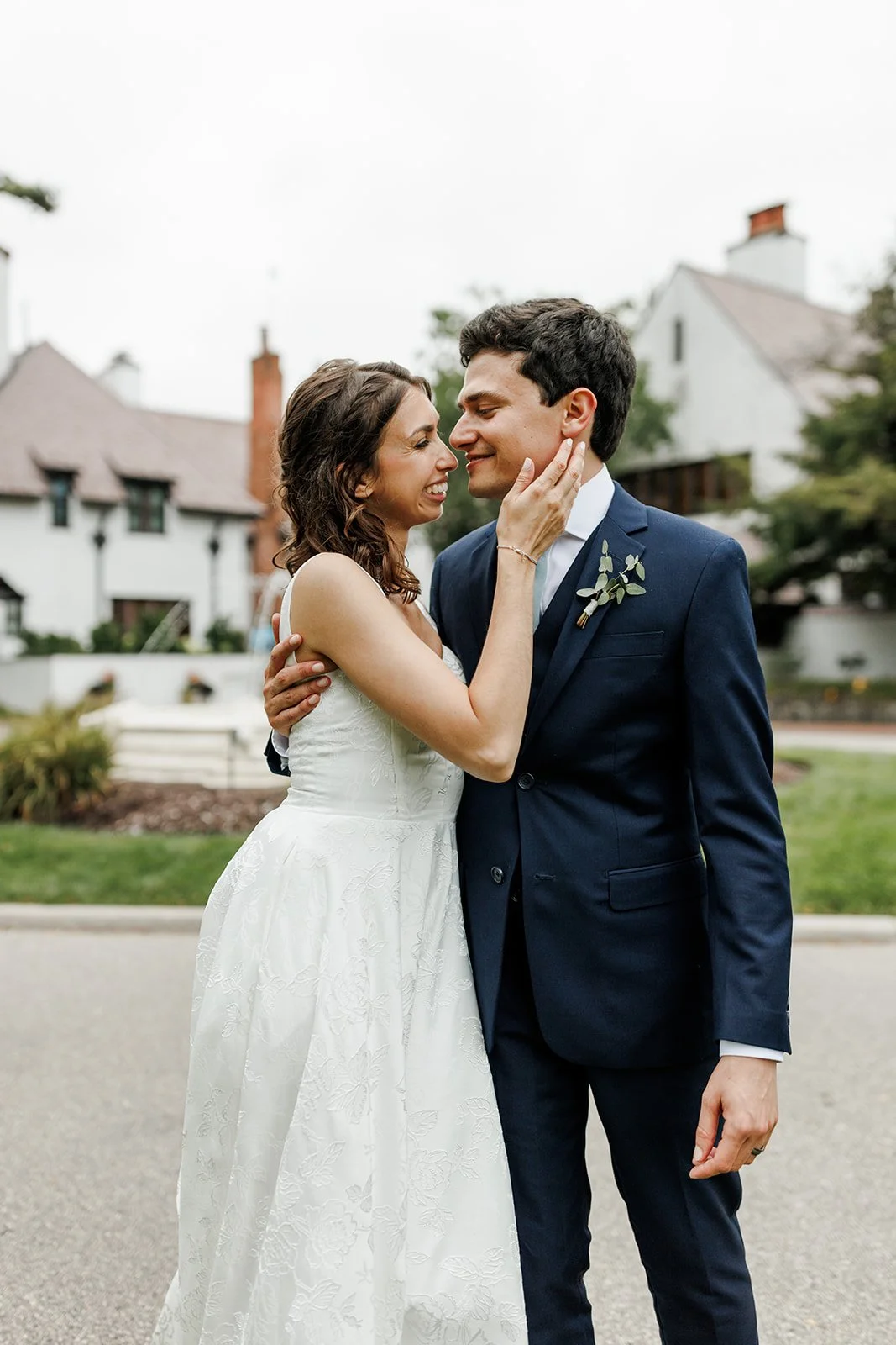 A bride and groom in wedding attire embrace outdoors, smiling at each other on a cloudy day.