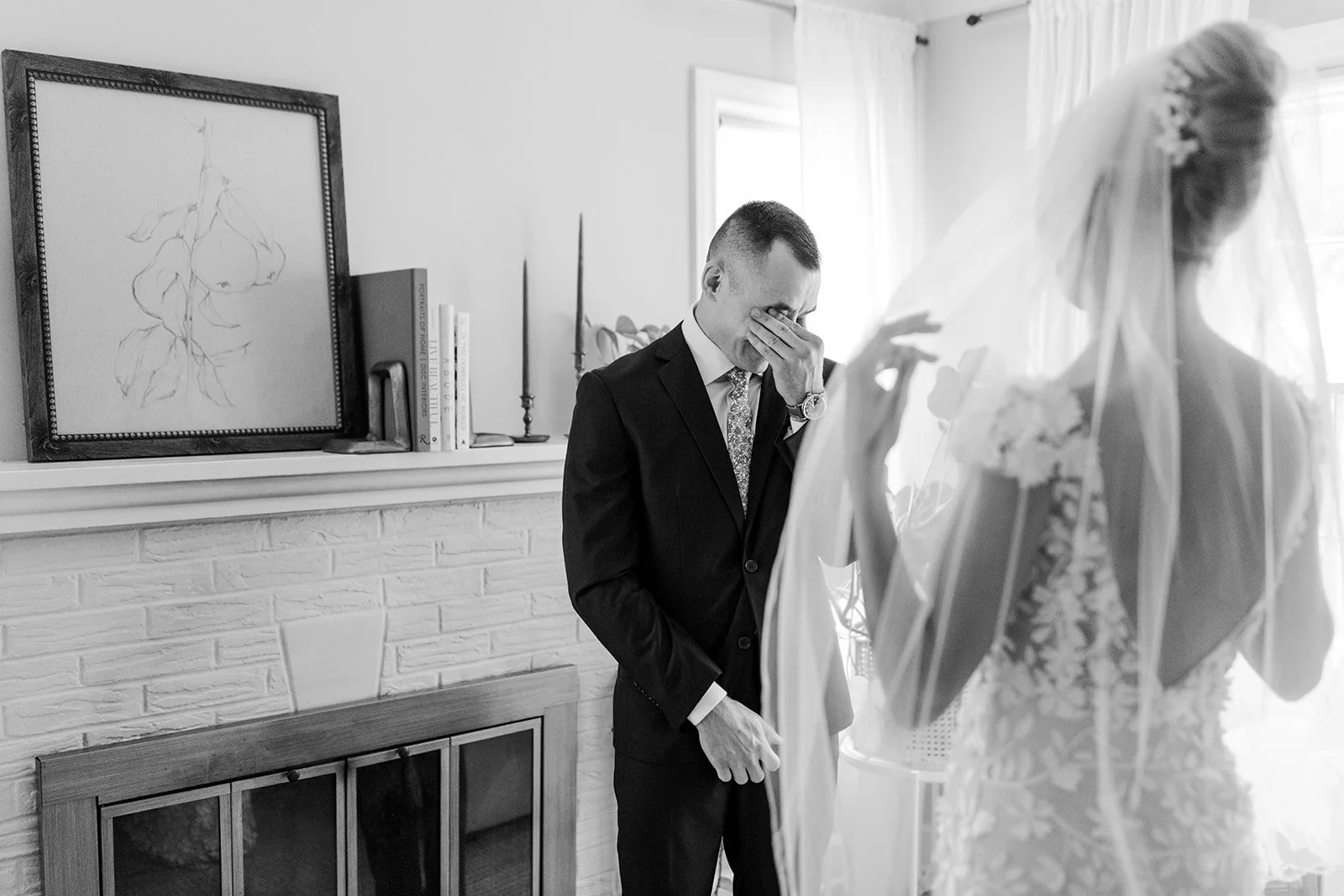 A bride in a wedding dress and veil stands inside, while a man in a suit, likely the groom, stands nearby with his hand covering his face in an emotional moment. The scene is in a cozy room with a brick fireplace, candles, and books on the mantle, an