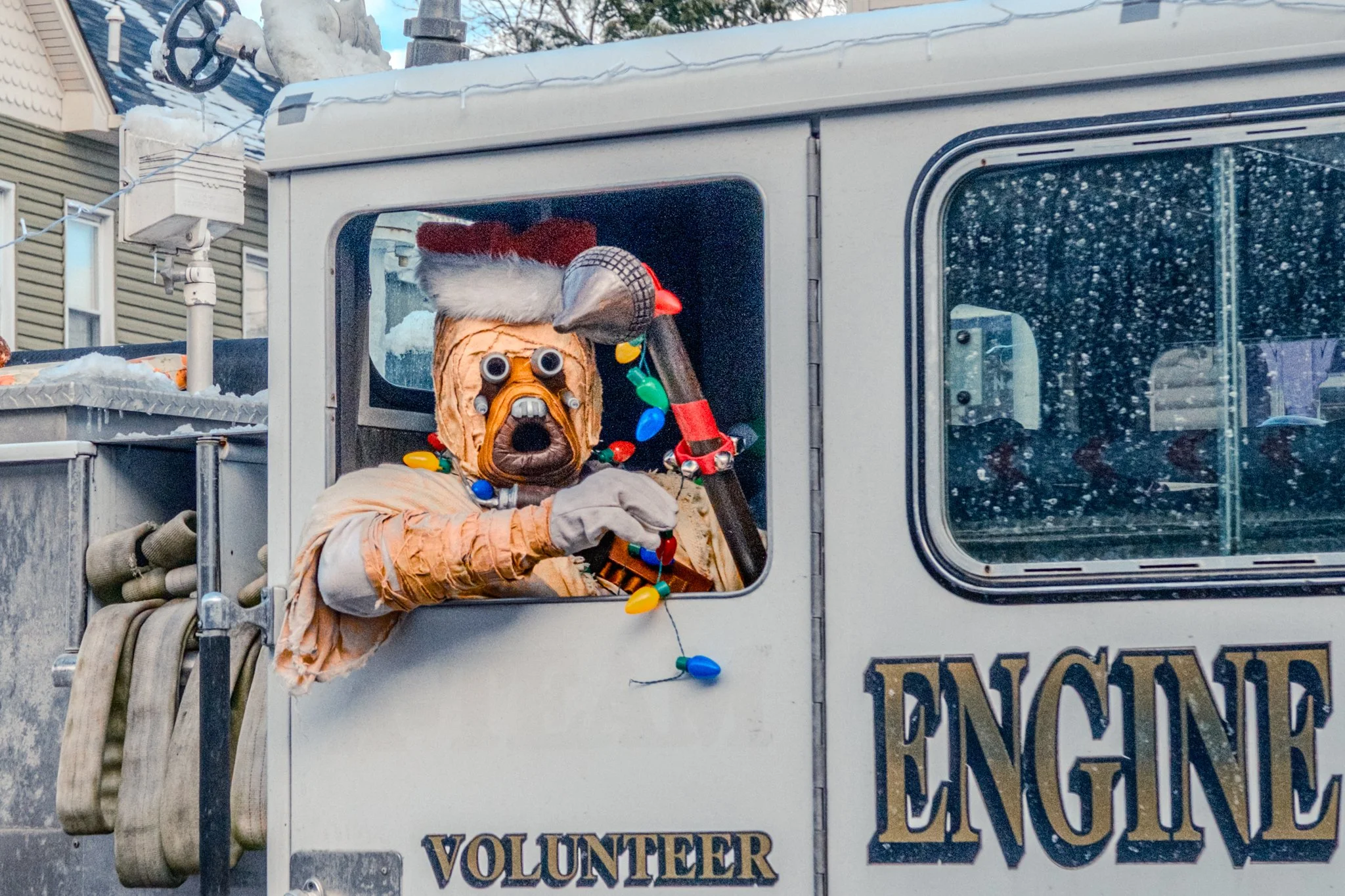Person dressed as an elf in a costume featuring a mask with a surprised expression, wearing a Santa hat, leaning out of a decorated vehicle with the words 'Volunteer' and 'Engine' on the side during a winter event.