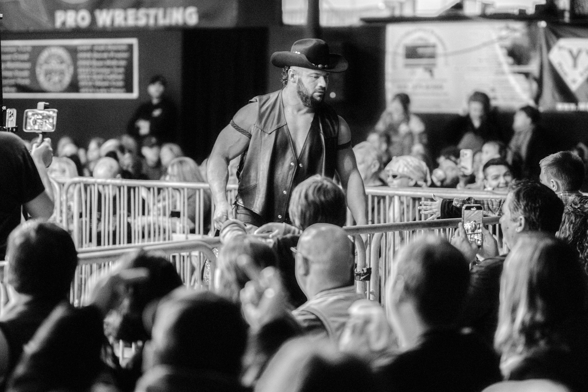 A black and white photo of a male wrestler in a leather vest and cowboy hat, standing behind a barricade, with an audience taking photos and watching at a wrestling event.
