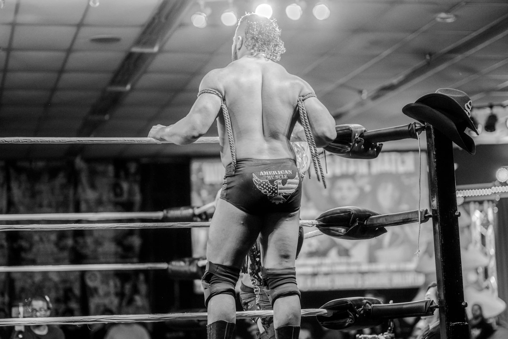 Professional wrestler with curly hair in a wrestling ring, facing away, wearing trunks with American-themed logo, surrounded by ropes, with a focused audience in the background.