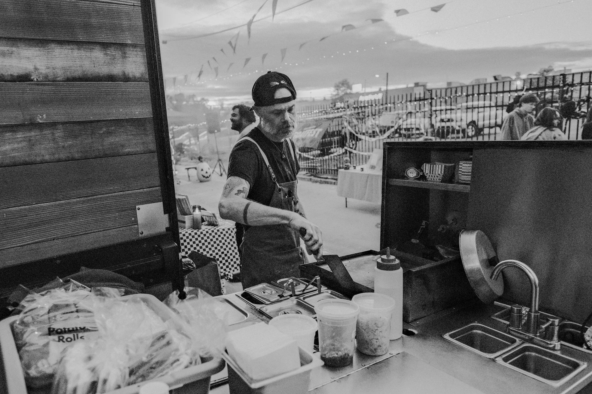 A man with tattoos, wearing a black baseball cap backward, preparing food at a food stand during an outdoor event with flags hanging overhead, a parking lot, and a few people in the background.