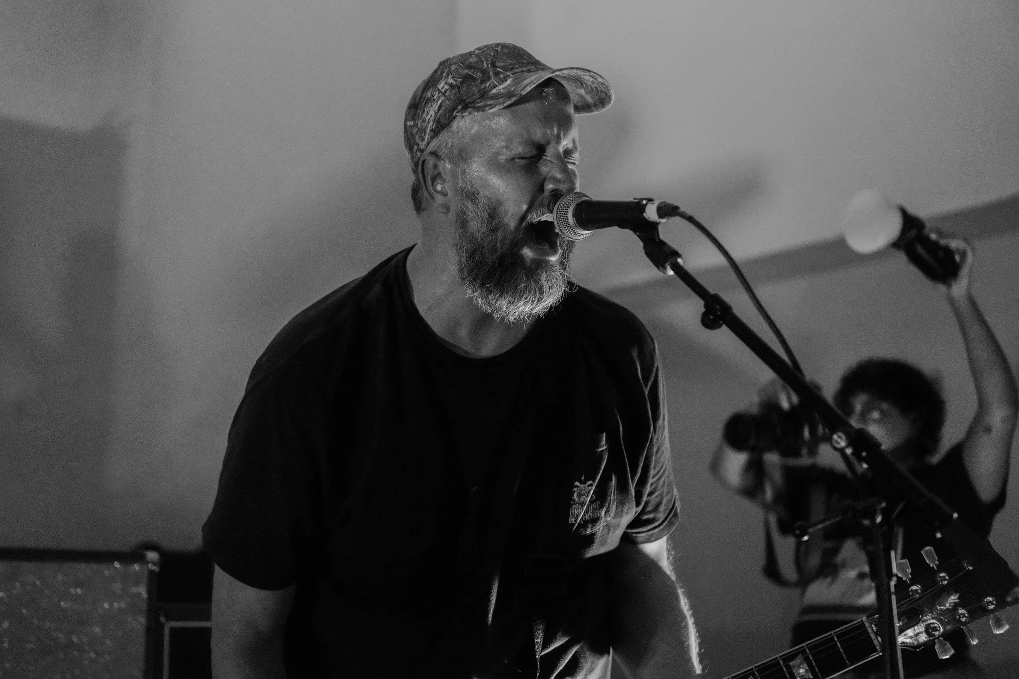 Black and white photo of a man with a beard singing into a microphone while playing an electric guitar, and a woman in the background taking photos with a camera.