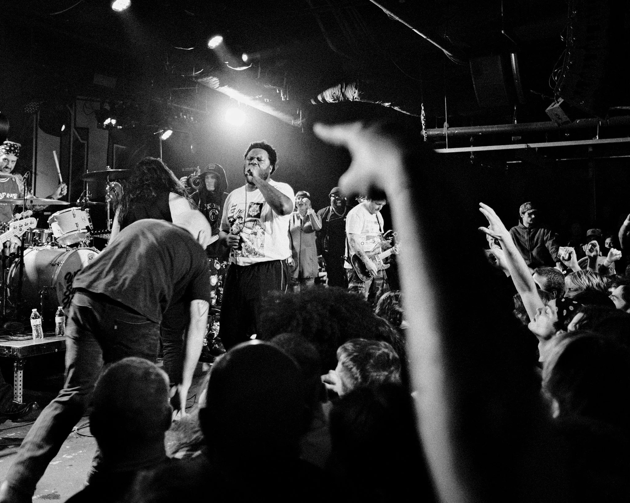 A black and white photo of a live concert with a band performing on stage and an audience cheering in front. Fans are raising their hands and dancing.