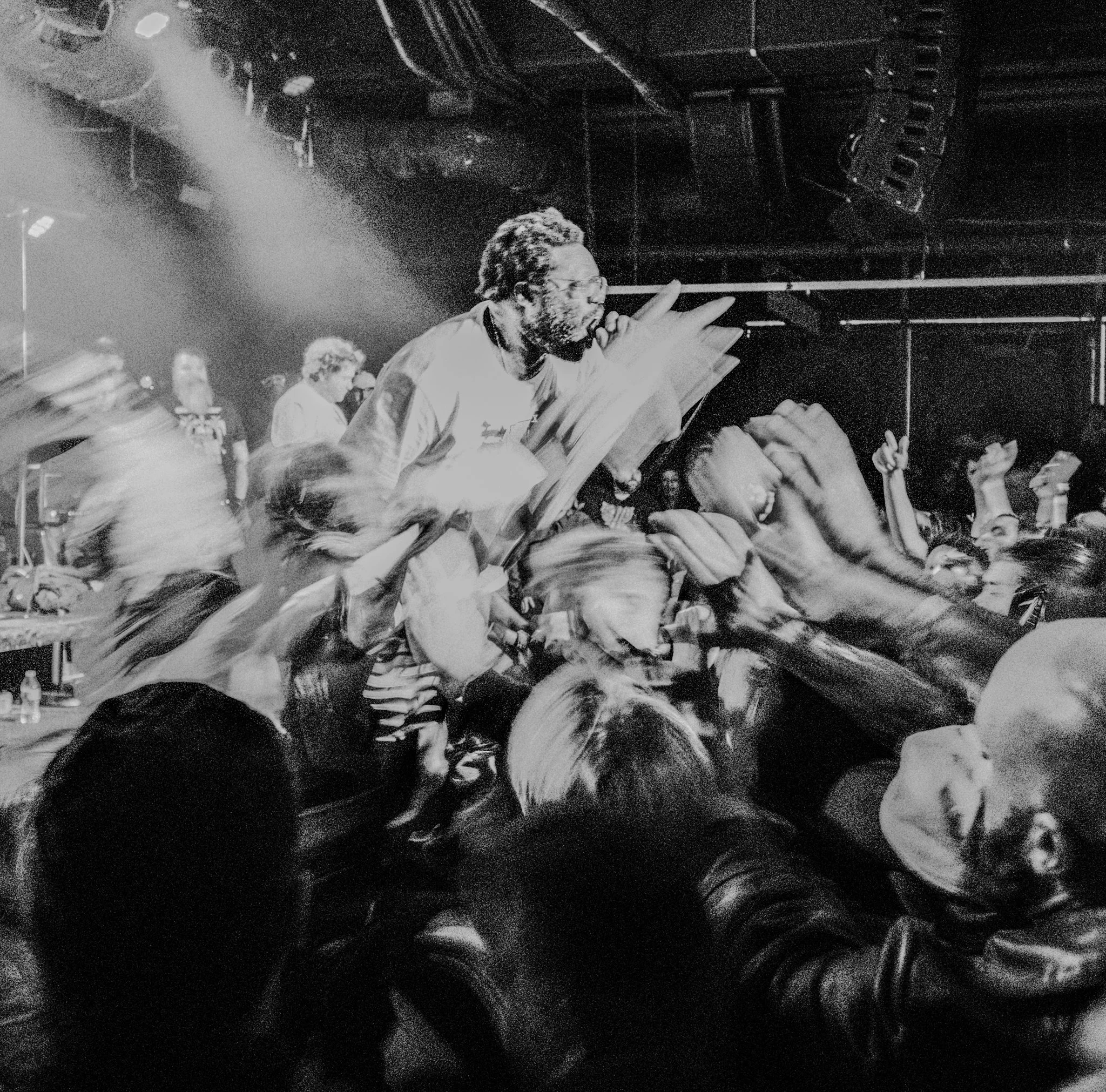 A black and white photo of a live concert showing a performer on stage with a microphone, engaging with the audience, who have their hands raised, in a dimly lit venue with stage lights.