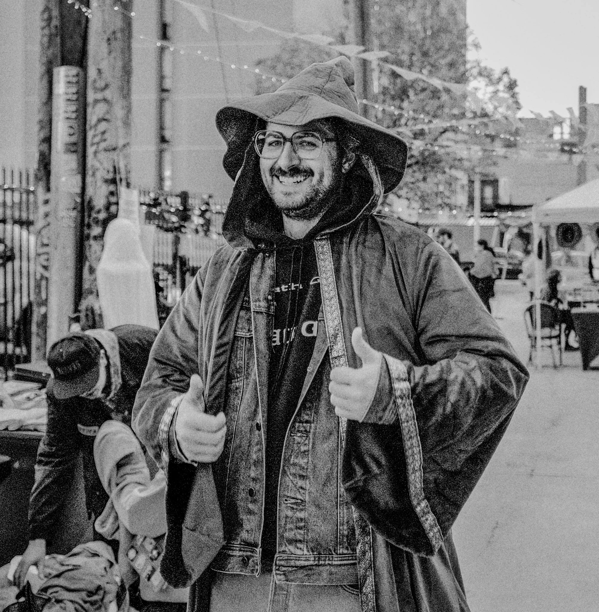 Man smiling outdoors with thumbs up, wearing a large hooded jacket and glasses, in an urban setting.