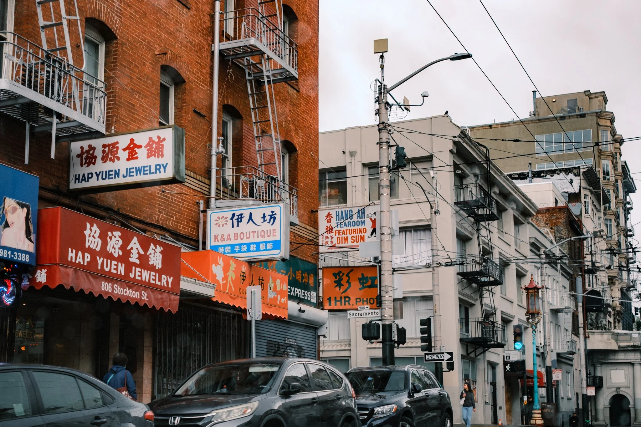 City street scene with multiple signs, buildings, cars, and pedestrians on Stockton Street in San Francisco, including jewelry and photo shops.