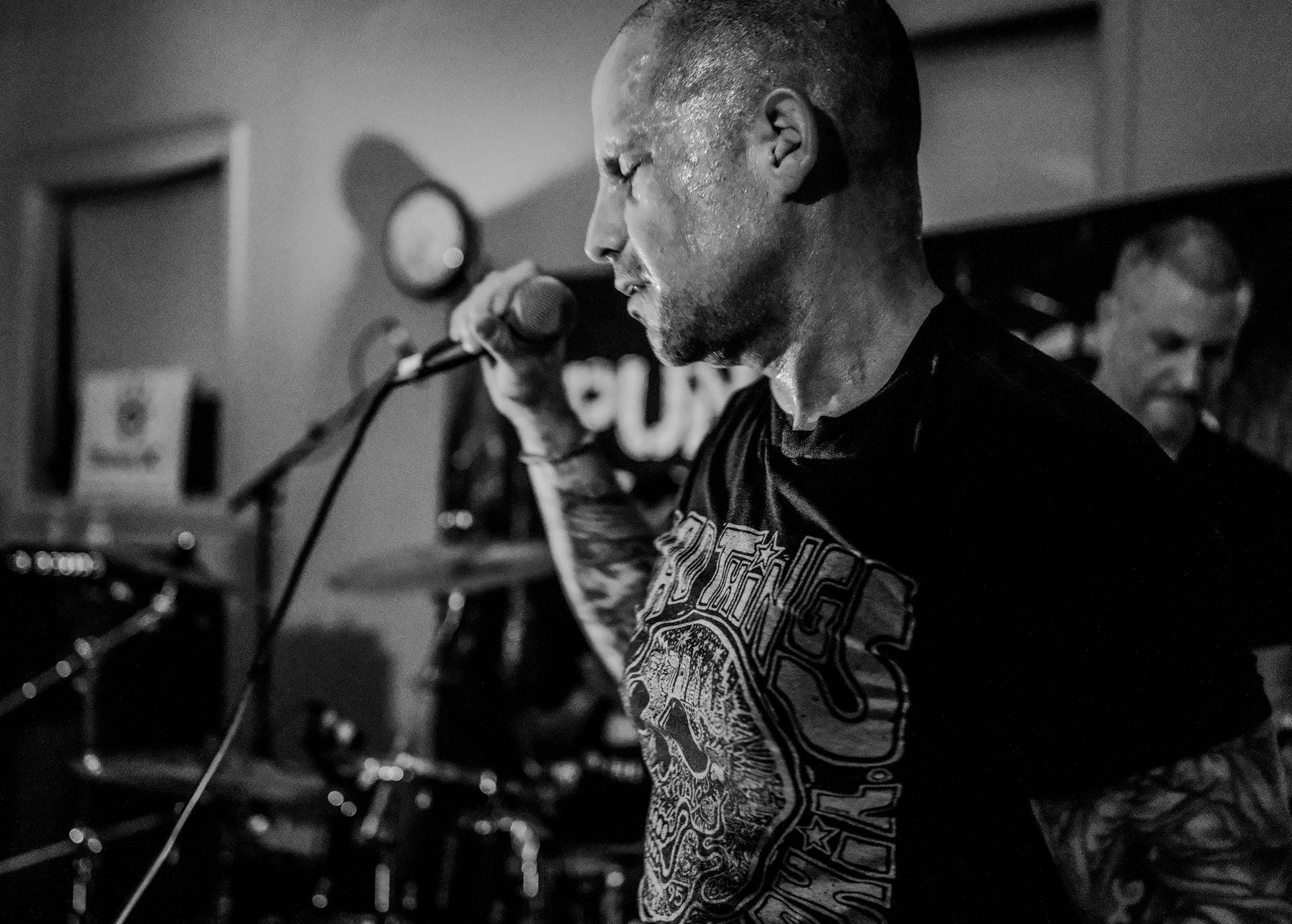 A man with tattoos on his arms, holding a microphone in front of his face, appears to be singing or shouting, with sweat on his face. A woman is seen in the background, smiling.