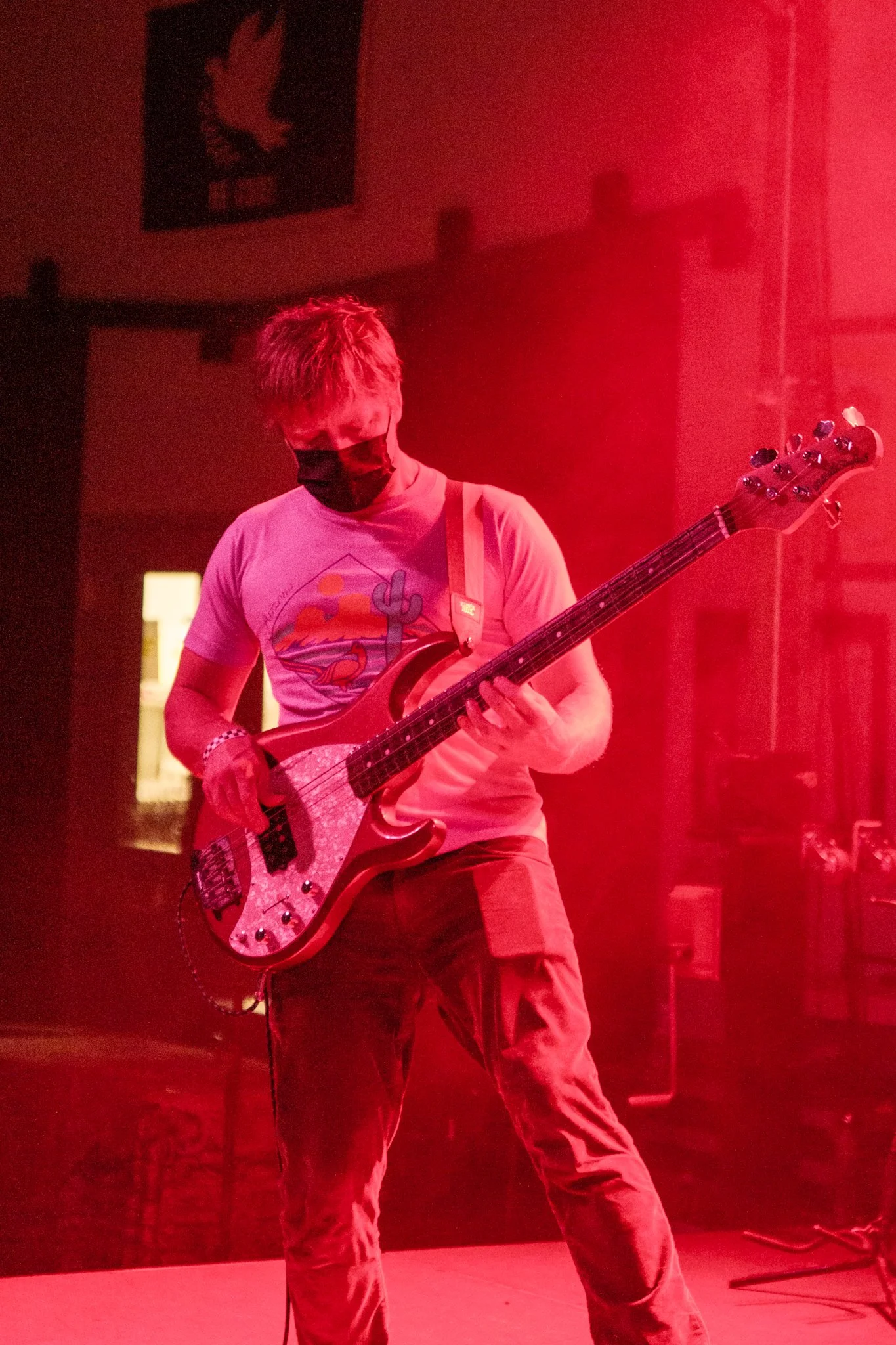 A person playing electric bass guitar on stage, wearing a black face mask, T-shirt with a colorful graphic, and cargo pants, with red lighting and a background banner featuring a bird logo.