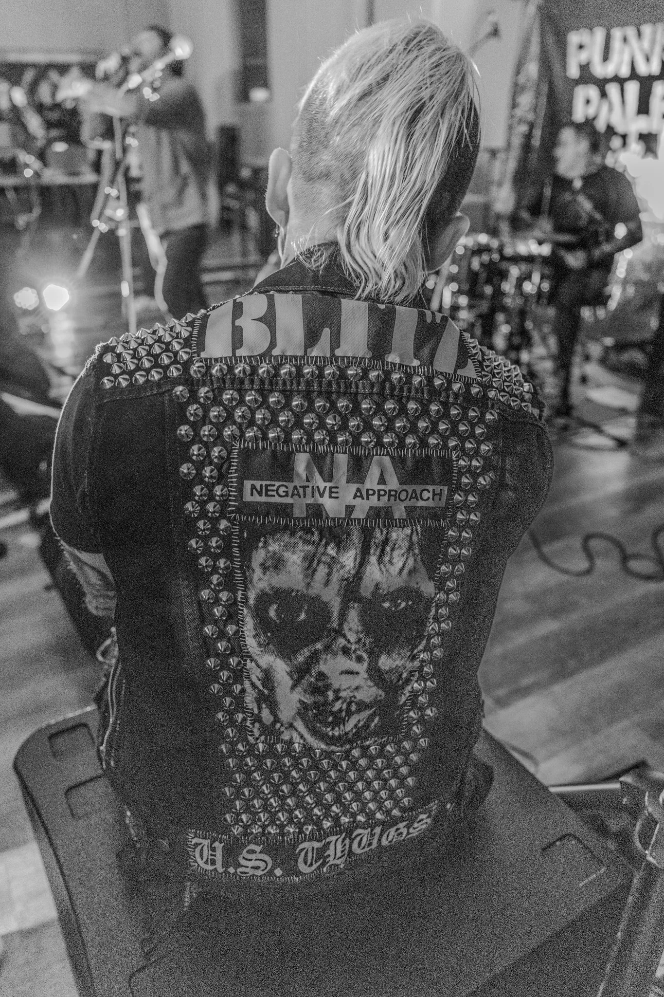 A black and white photo of a person with a punk hairstyle, wearing a studded denim vest with patches, sitting on a stool facing a band performing on stage.