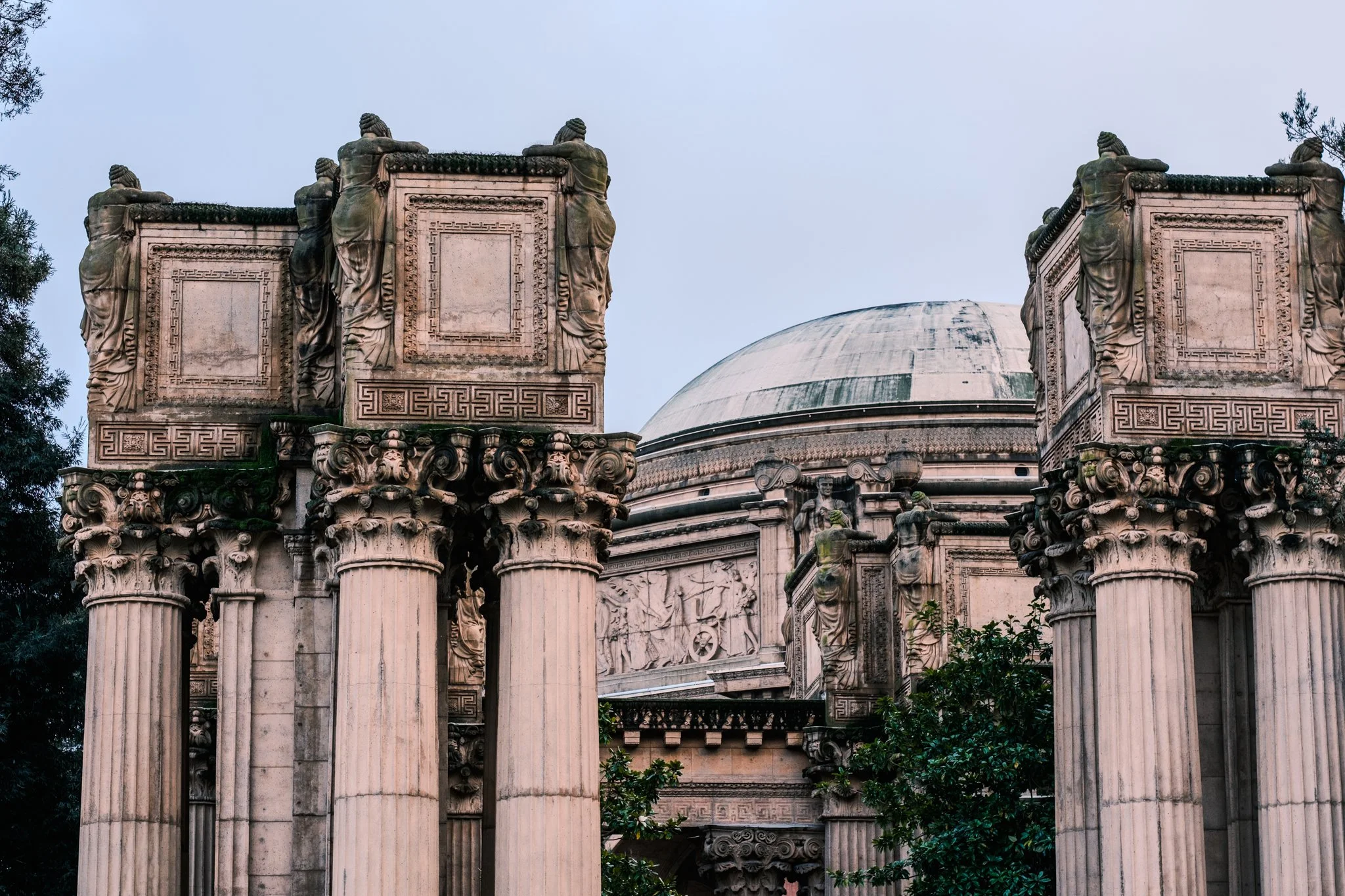 Ancient ruins with tall columns and a domed structure, decorated with sculptures and intricate carvings, surrounded by greenery.