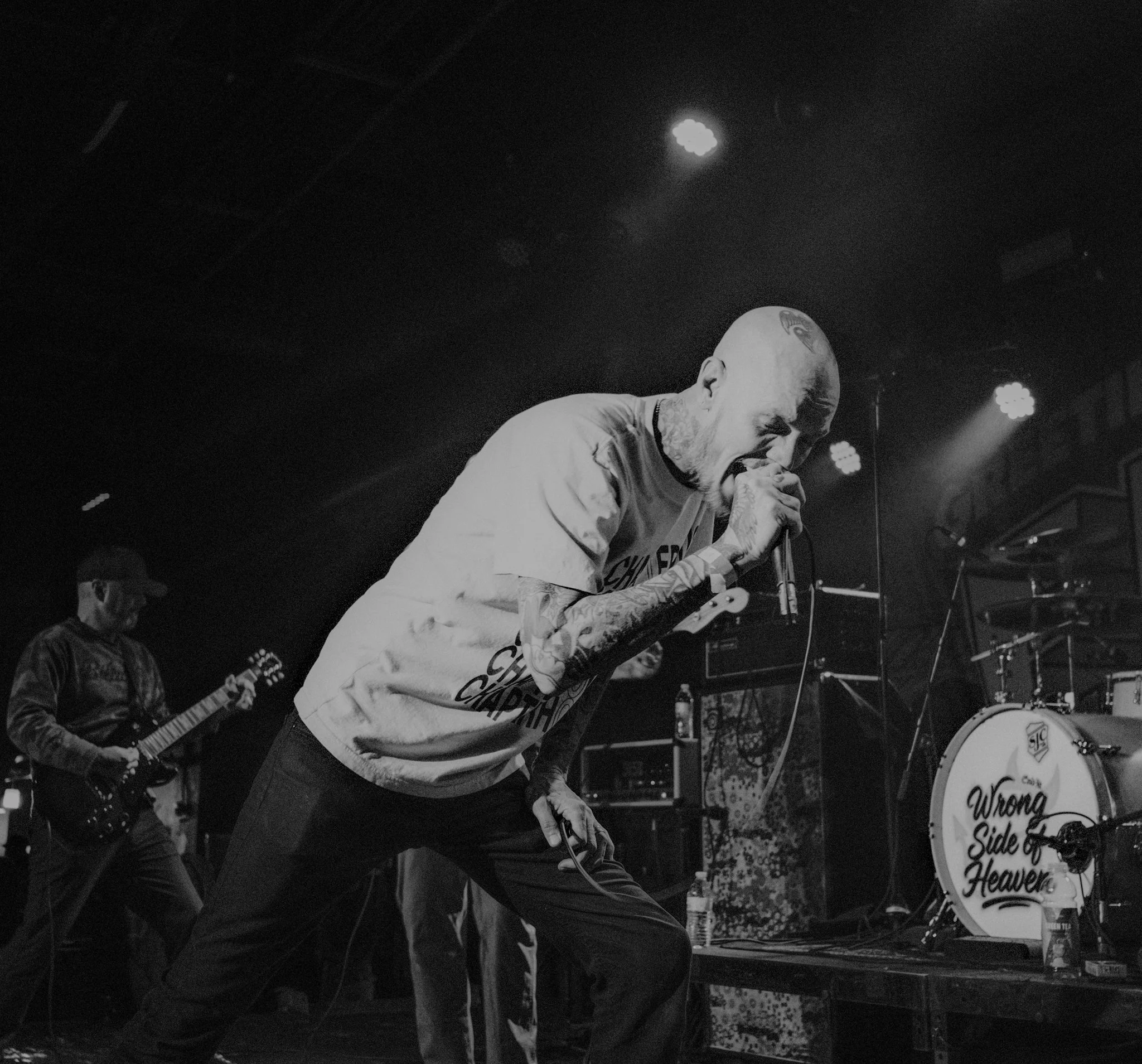 A black and white photo of a band performing on stage with the lead singer passionately singing into a microphone, tattoos visible on his arms, with a guitarist playing in the background and a drum set labeled "Wrong Side of Heaven" on stage.