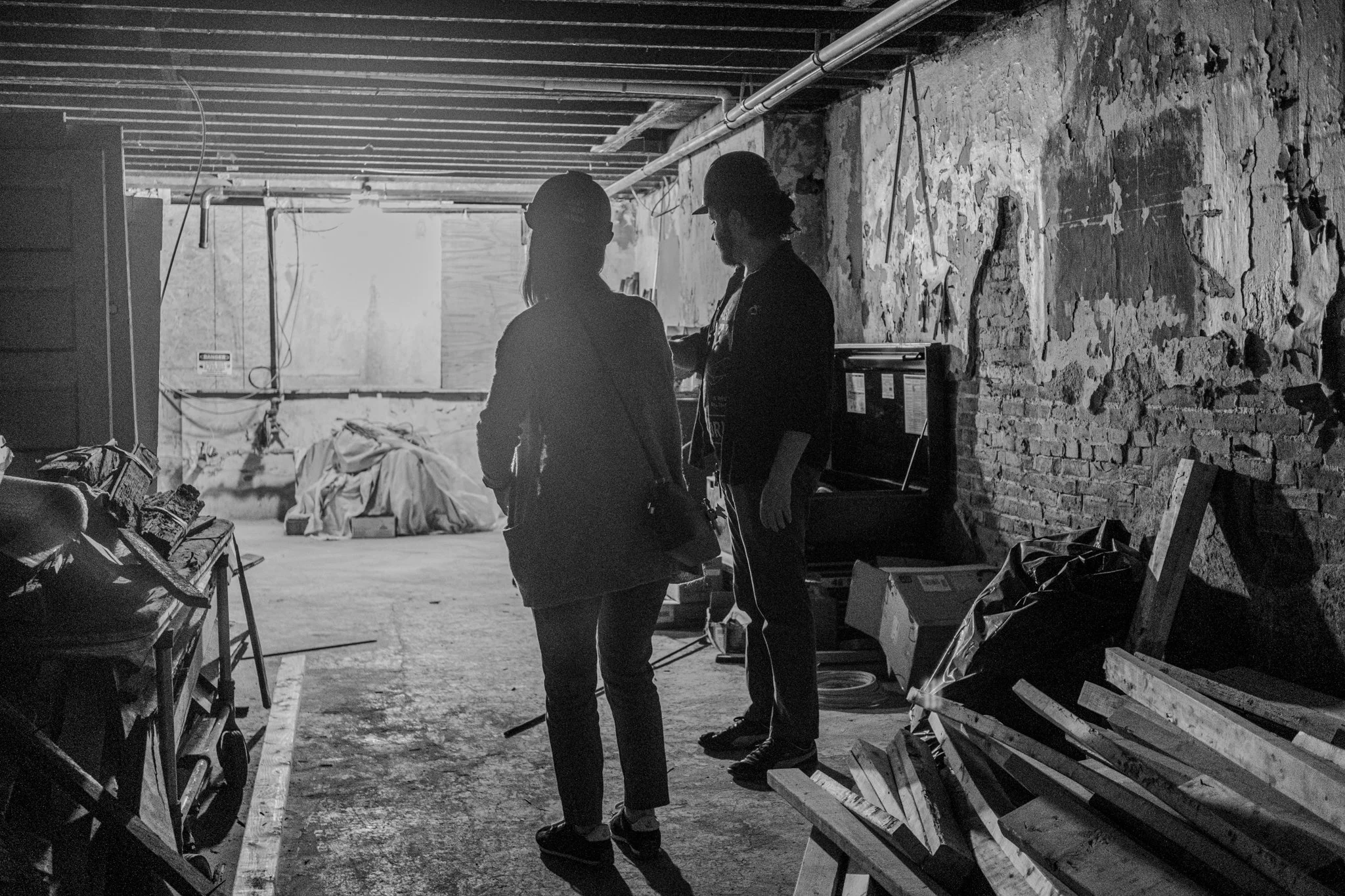 Two people standing inside an unfinished or abandoned building with exposed brick walls and construction debris, engaged in conversation.