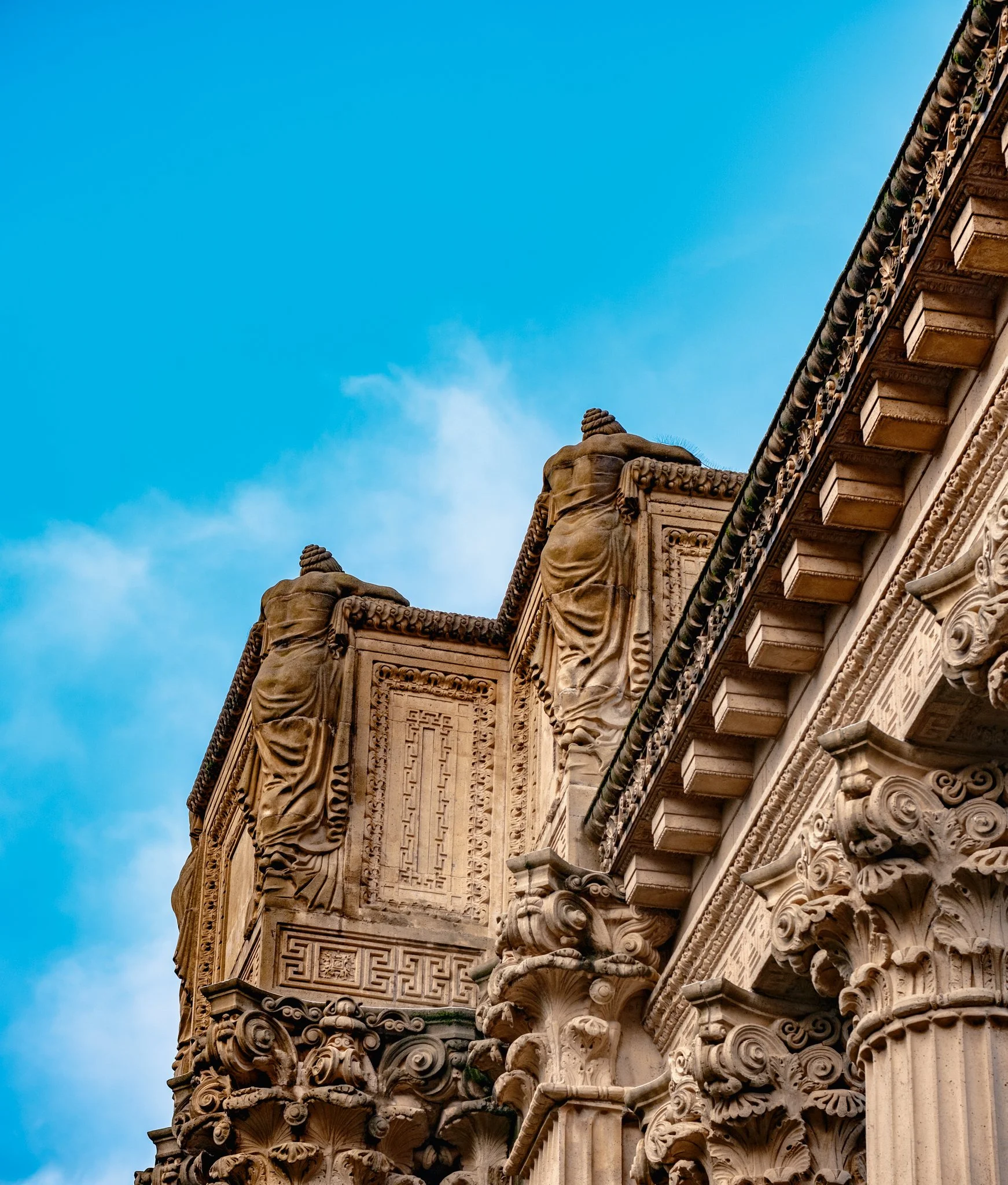 Close-up of ornate ancient Greek or Roman stone architectural details with columns and sculptures, against a clear blue sky.