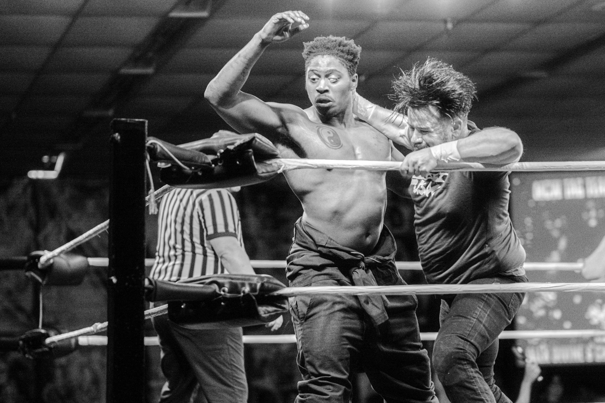 Two men wrestling in a ring, one is shirtless and the other wears a t-shirt, with a referee in the background.