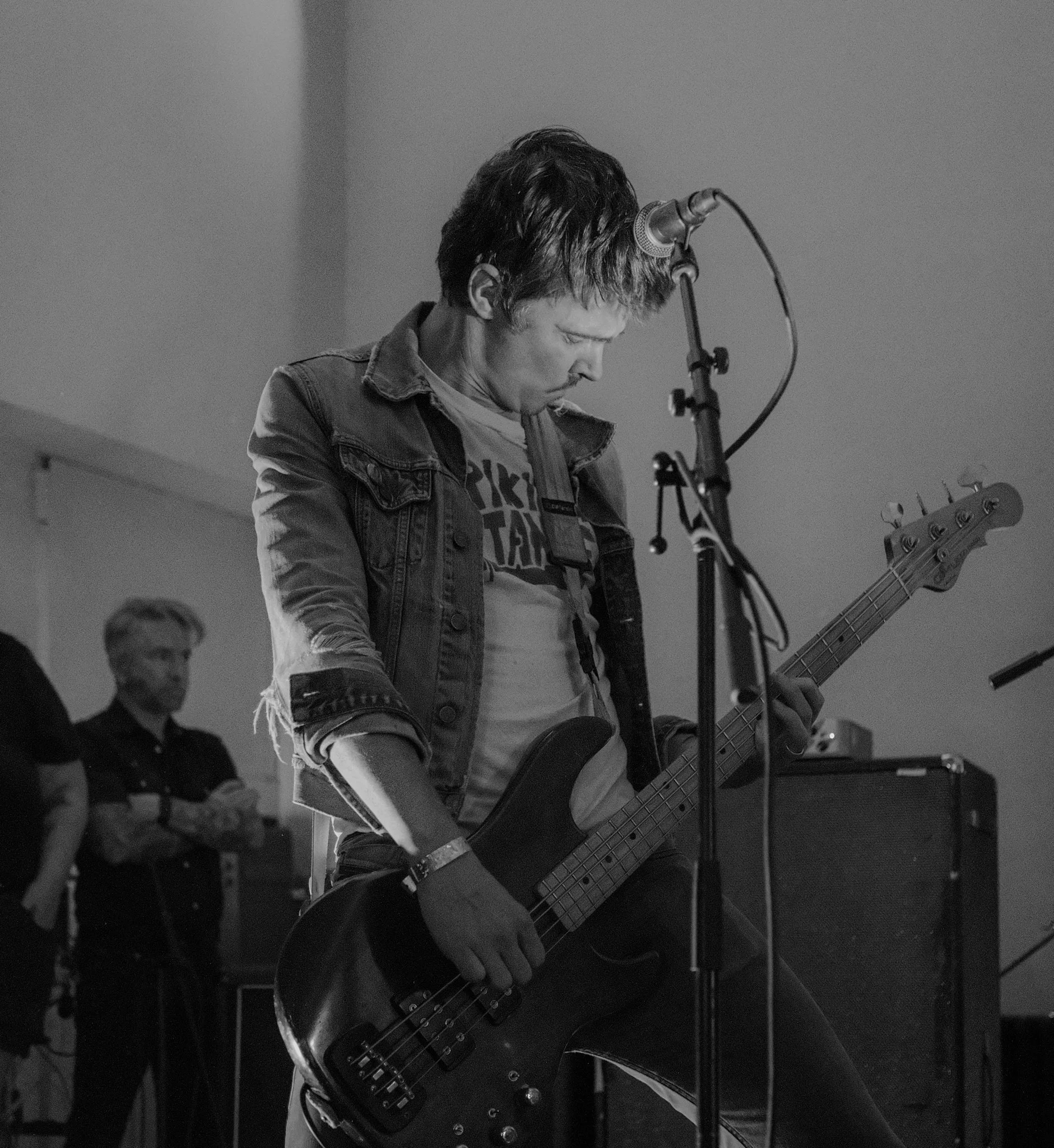 A person playing an electric bass guitar on stage with a microphone, wearing a denim jacket and a t-shirt, while another person looks on in the background.