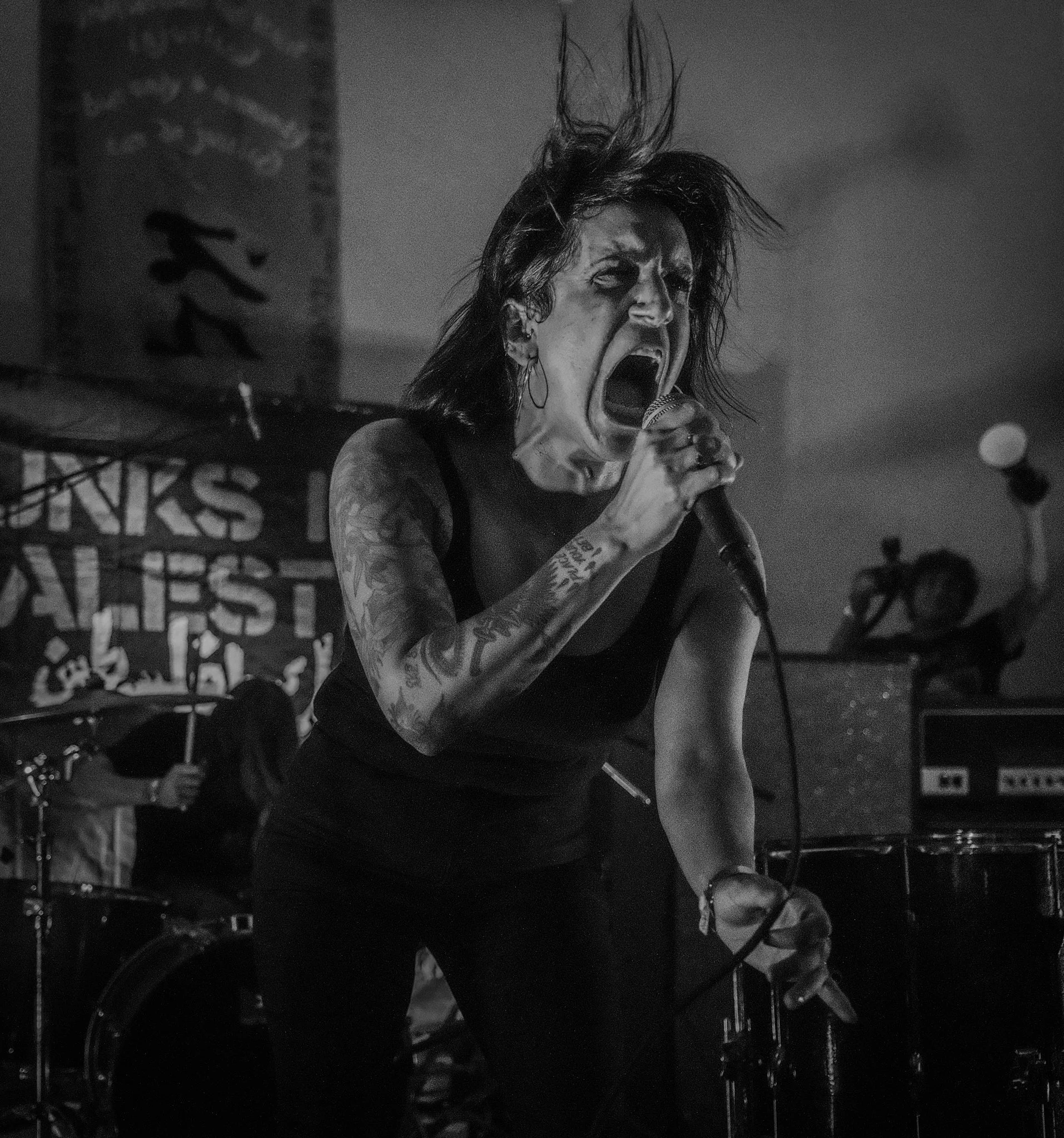 A female singer passionately performing on stage, gripping a microphone with her mouth open wide. She has tattoos on her arms and dark, wild hair.