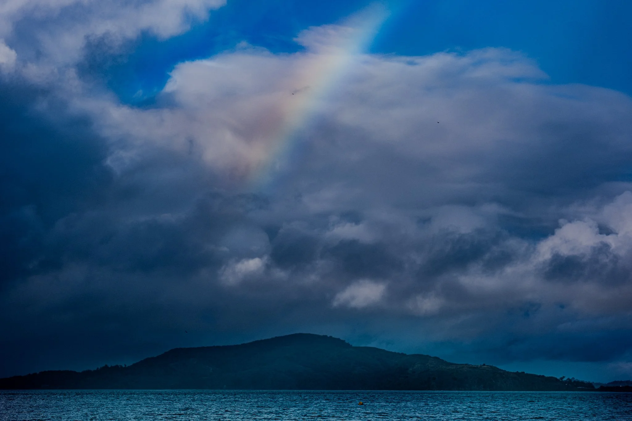 Cloudy sky over a body of water with a faint rainbow and a distant mountain or island.
