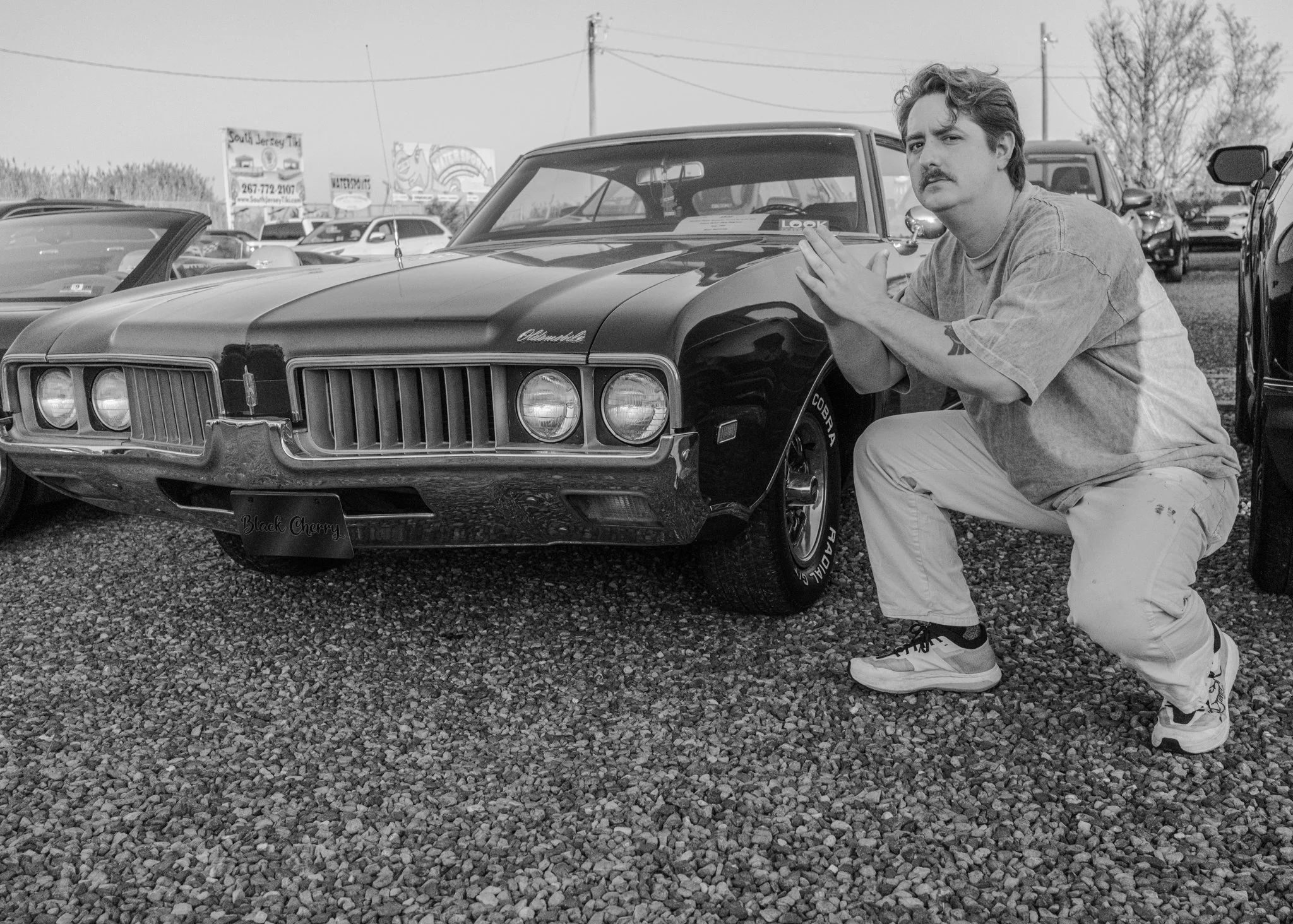 A man crouching next to a vintage black Chevrolet muscle car at a car show, with other cars and signboards in the background.