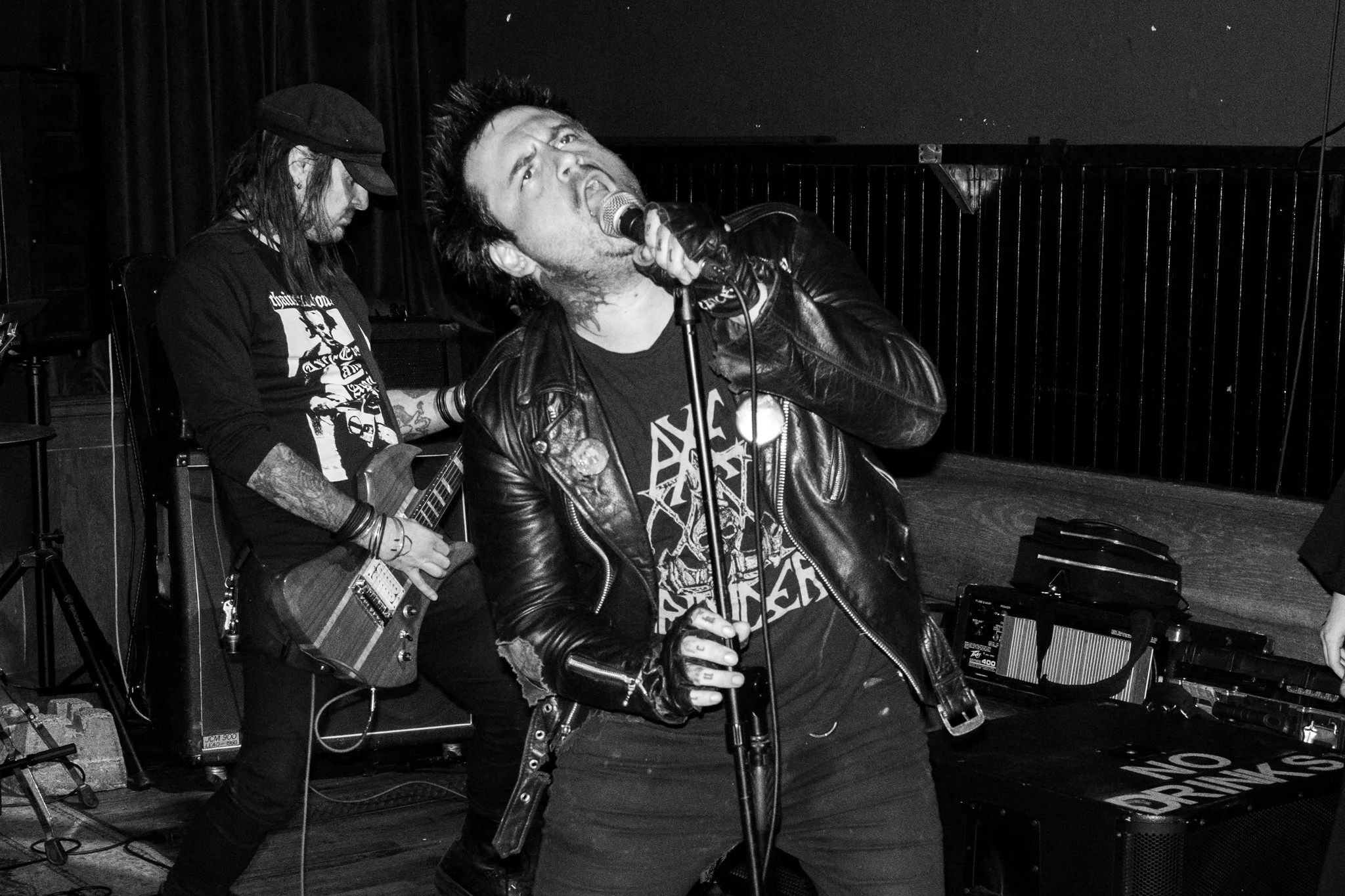 Black and white photo of a lead singer and guitarist performing at a concert. The singer is wearing a leather jacket and singing into a microphone, while the guitarist, wearing a hat, is playing an electric guitar in the background.
